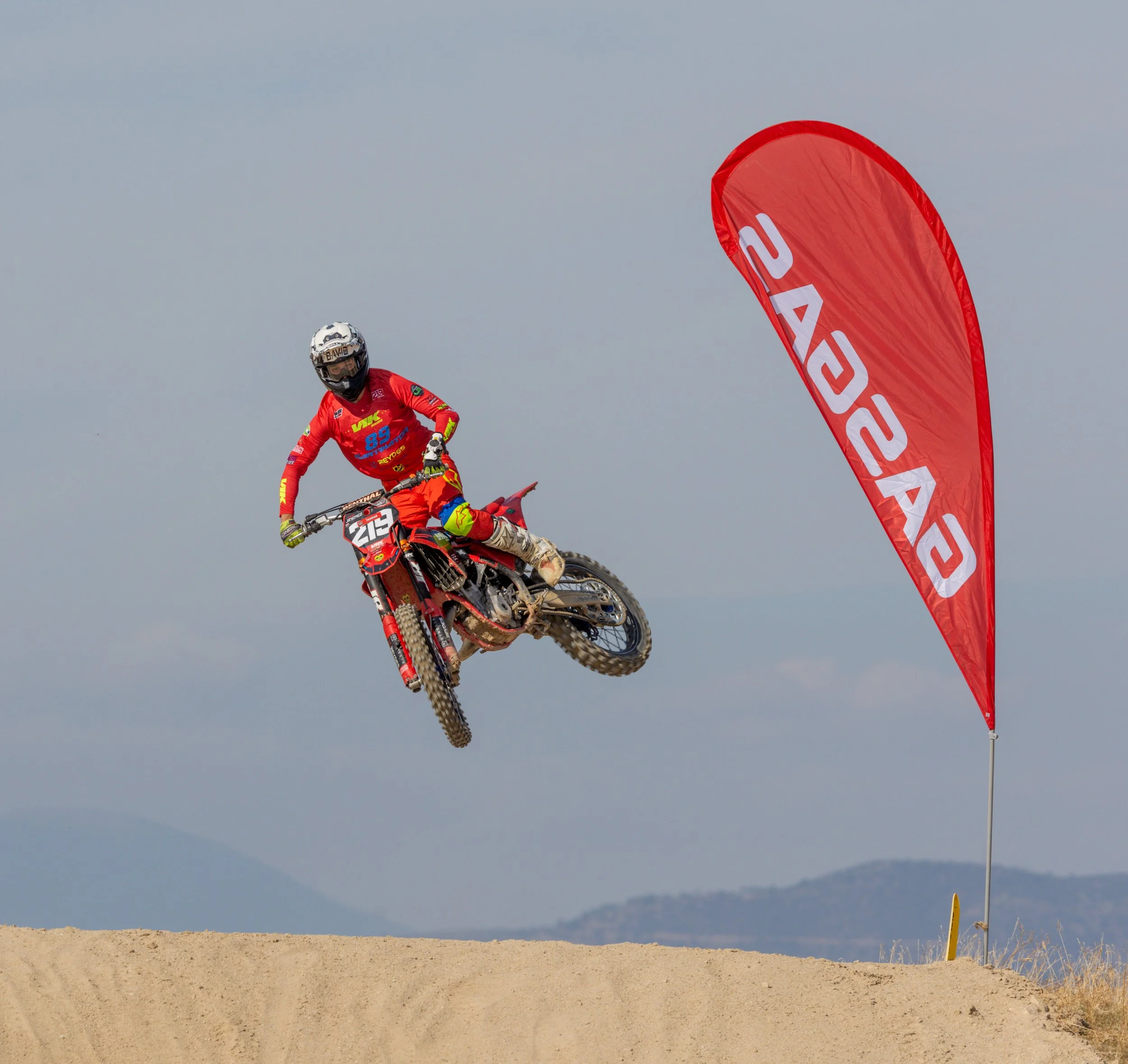 Motociclista en pleno salto en una carrera, con bandera roja que dice 'EASG' en el fondo de un cielo nublado.
