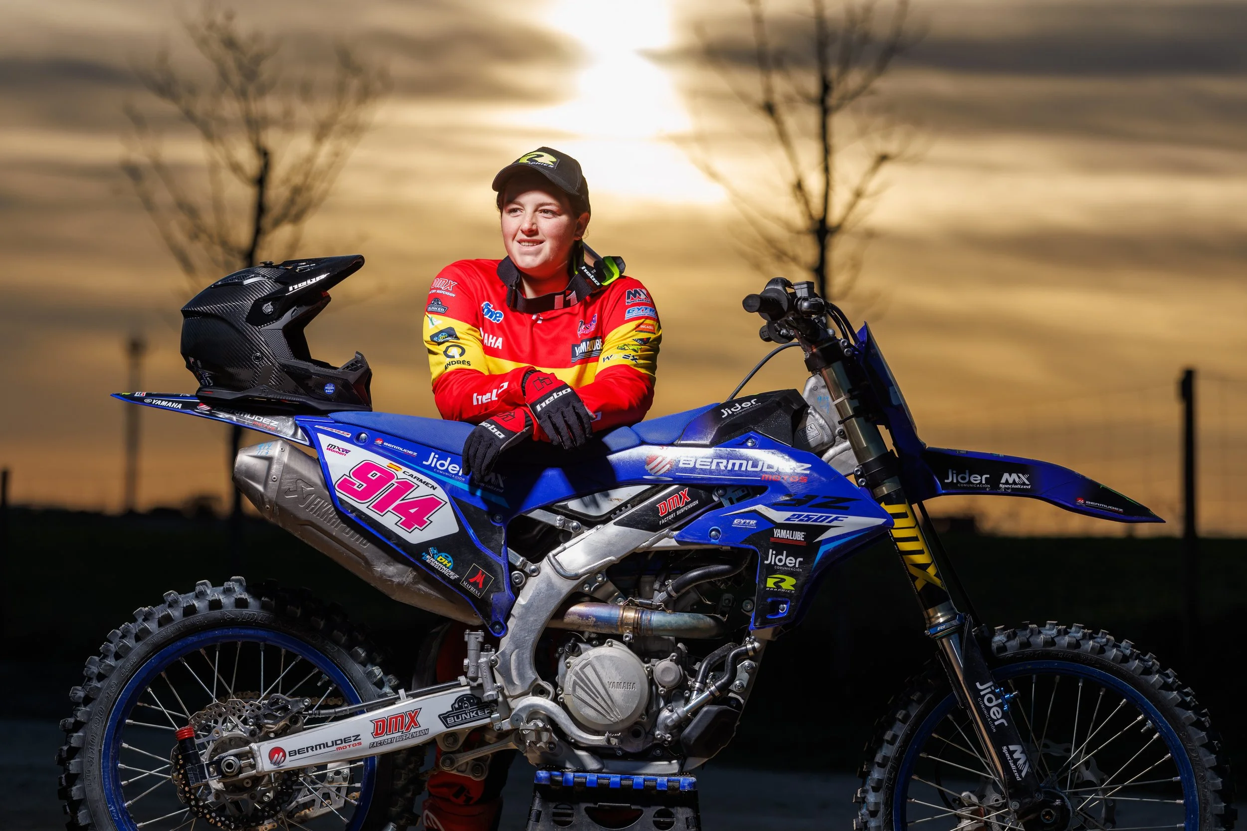 Una piloto de motocross sonriendo mientras posa con su motocicleta azul y varios patrocinadores visibles, en un atardecer con árboles en el fondo.