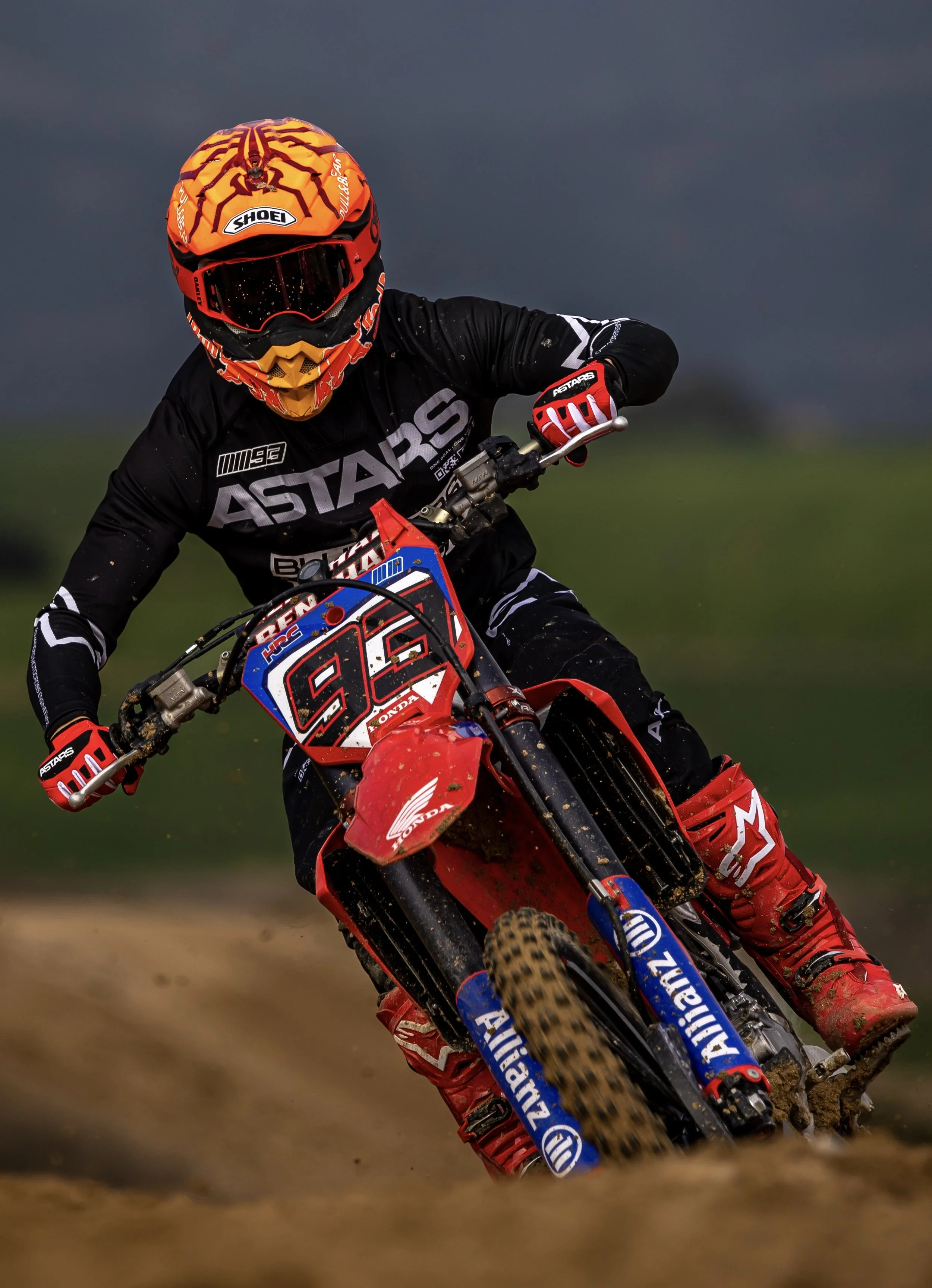 Persona en una motocicleta de motocross en un circuito, con casco naranja con rojo y gafas oscuras, vestido con ropa negra y botas rojas y negras, en movimiento y lanzando tierra.