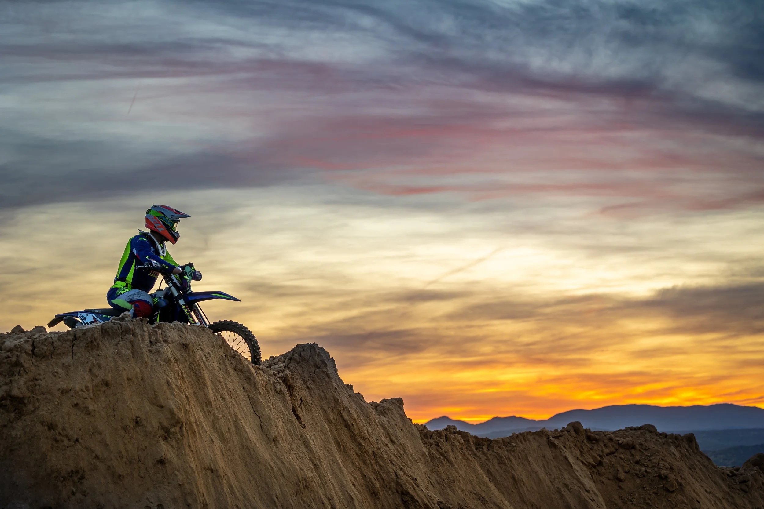 Un motociclista en equipo de protección, sentado en una motocicleta sobre un terreno rocoso al atardecer, con un cielo colorido y montañas al fondo.
