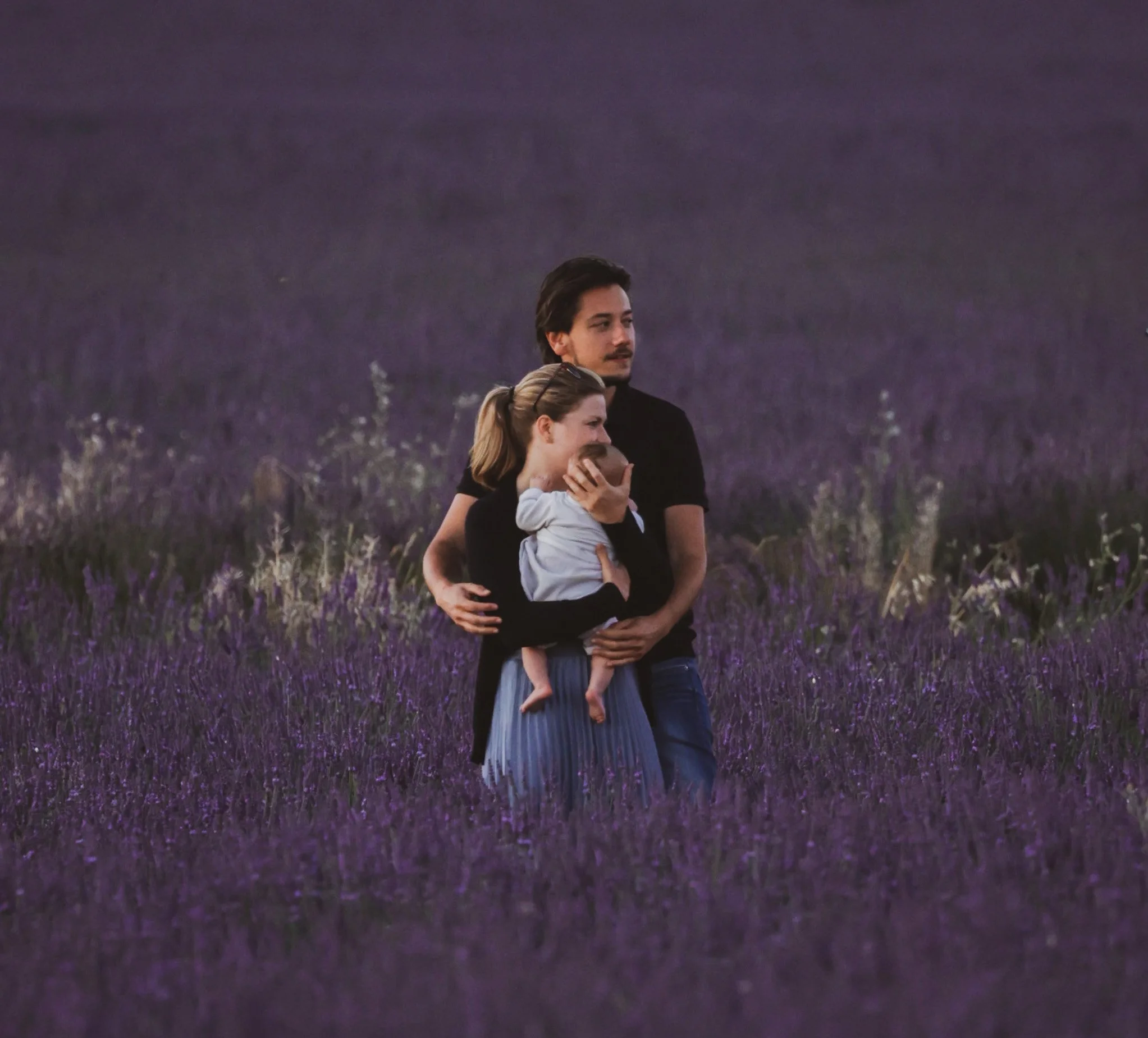 Una familia de tres en un campo de lavanda, con un hombre, una mujer y un niño pequeño. La mujer sostiene al niño y el hombre la abraza desde atrás.