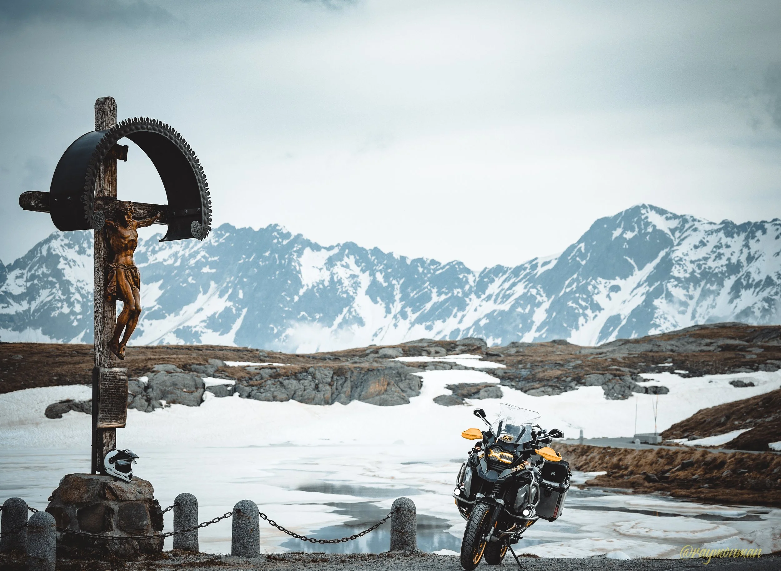 Una motocicleta negra con detalles amarillos estacionada en un paisaje montañoso con nieve y un lago congelado. A la izquierda, un crucifijo con una figura de Jesús crucificado y un casco de motocicleta en la base, rodeado por una cadena sujetada a p
