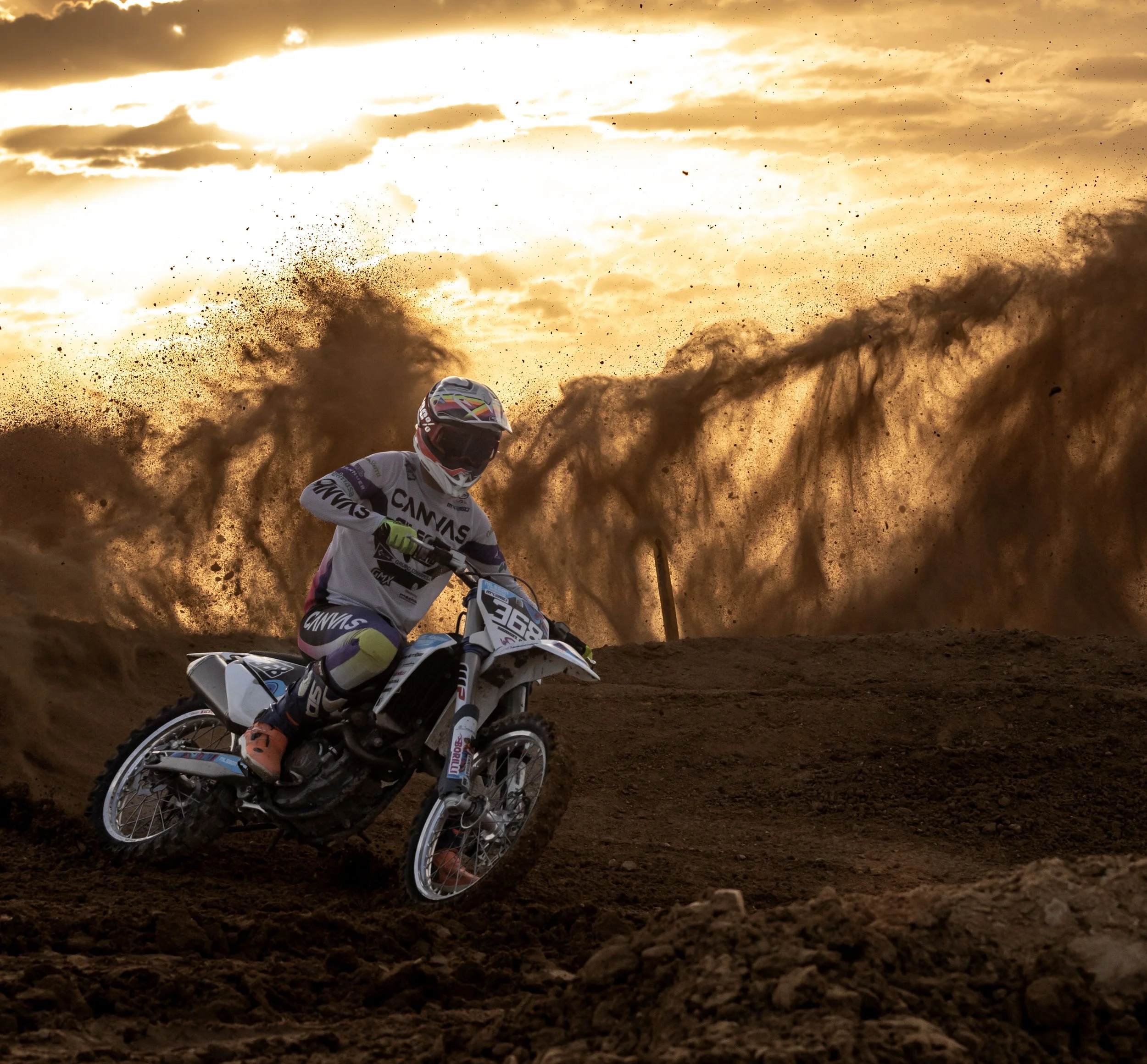 Un piloto de motocross en una pista de tierra, rodeado de polvo y arena, con un atardecer dorado en el cielo