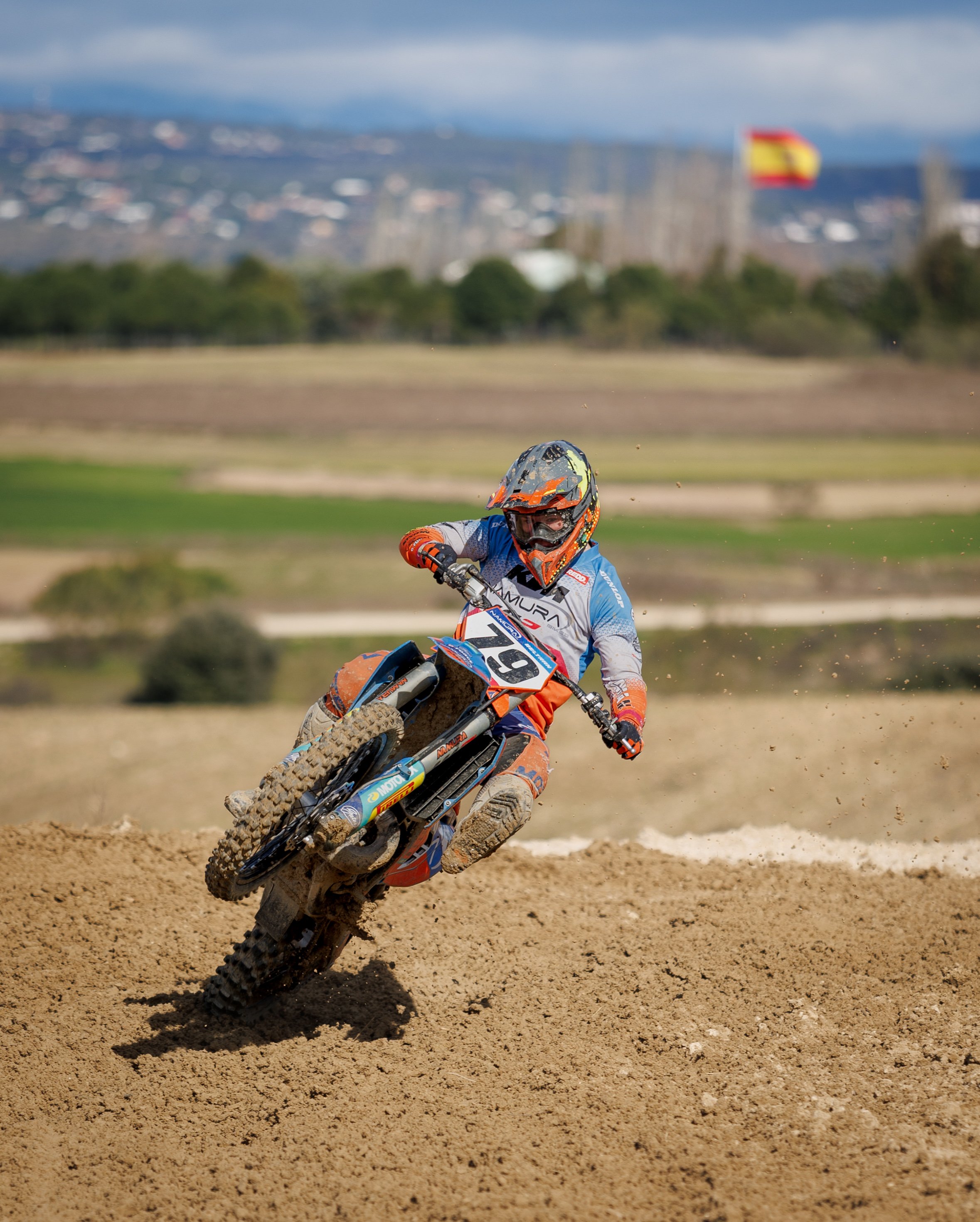 Un piloto de motocross en una carrera, vestido con equipo de protección naranja y azul manejando una motocicleta en una pista de tierra.