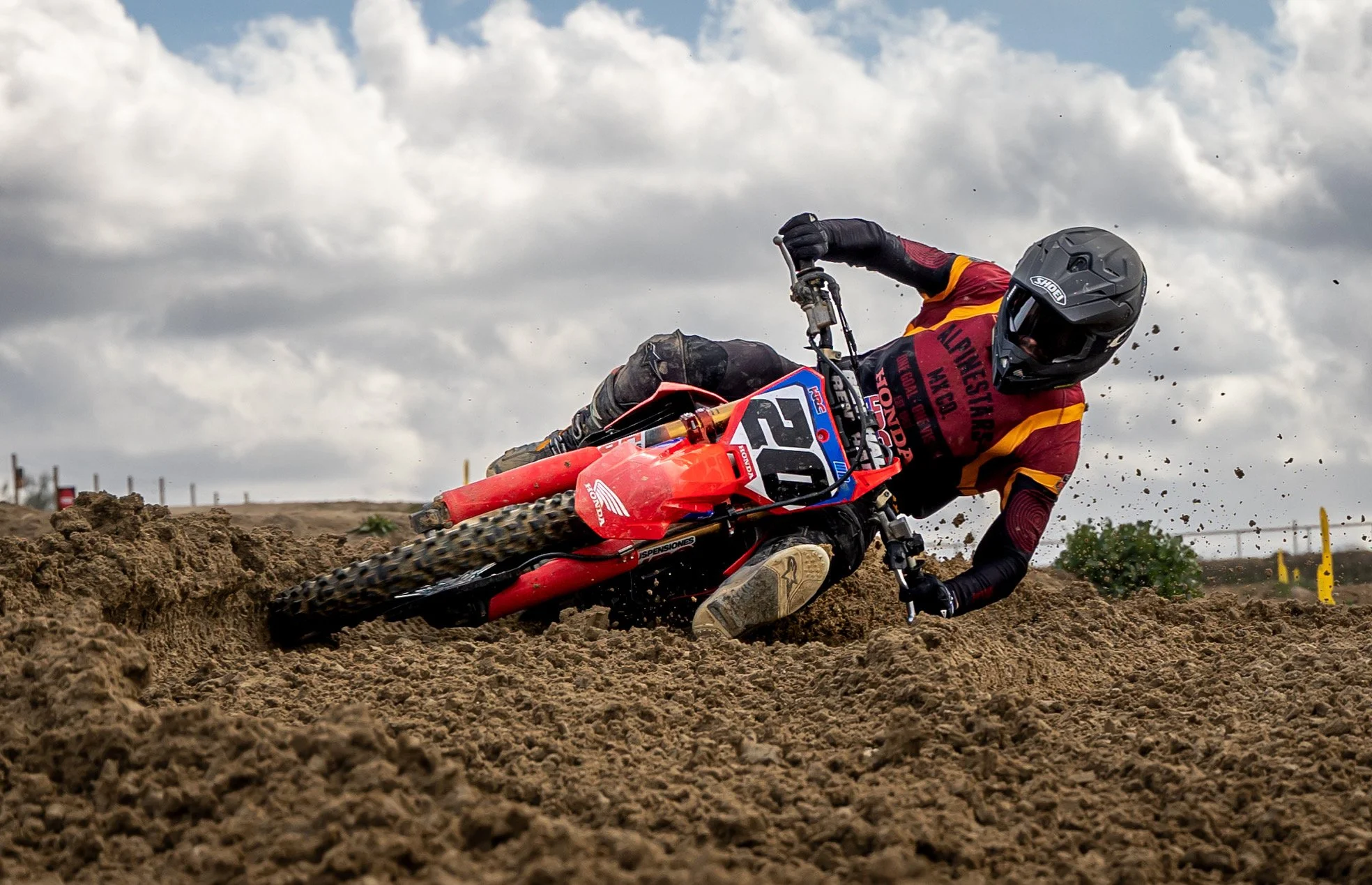 Un motociclista en una carrera de motocross en un terreno terroso, vestido con casco, ropas negras y un jersey rojo con amarillo, montando una moto roja con el número 26.