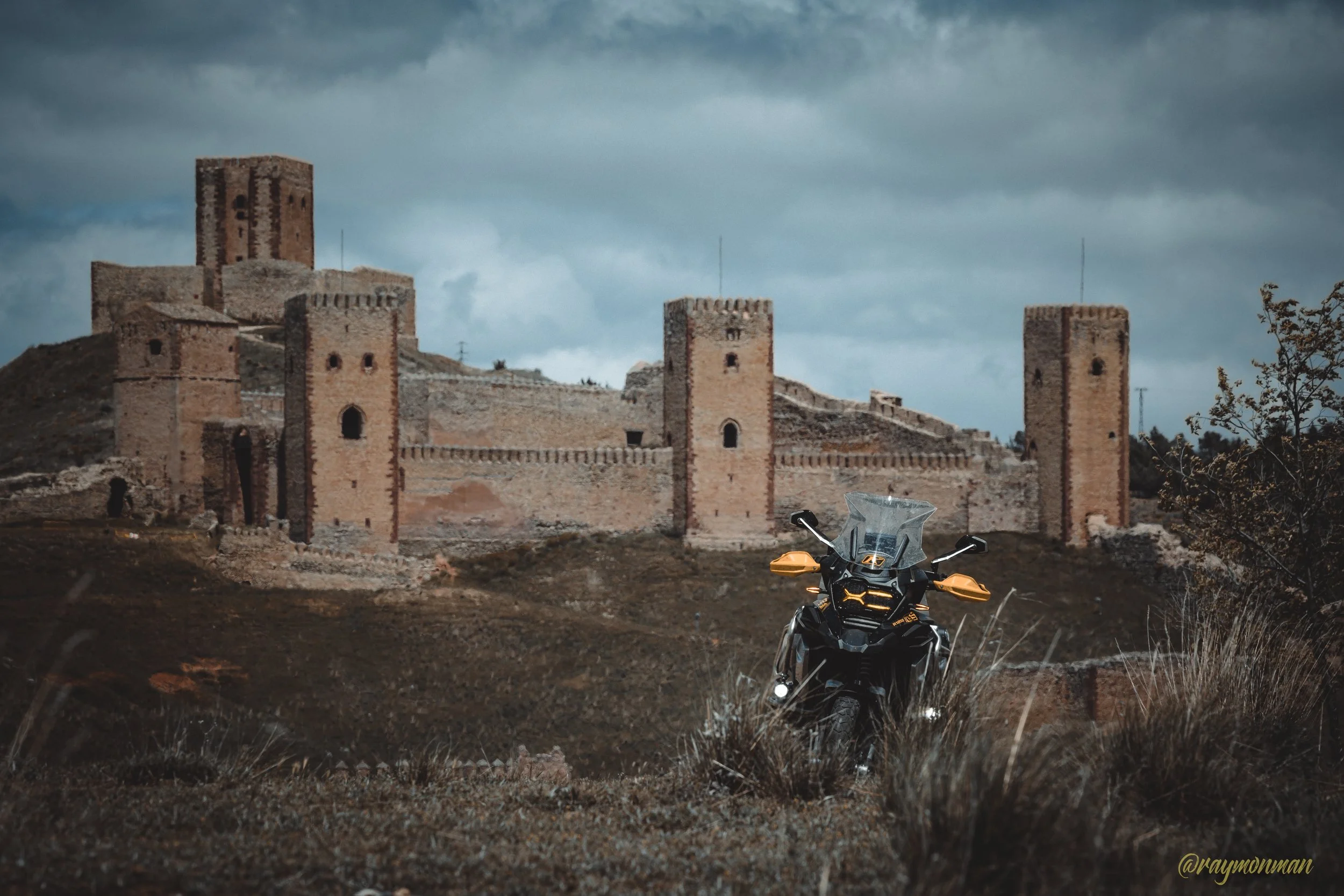 Una motocicleta negra con detalles dorados en primer plano, con un castillo de piedra en ruinas en el fondo, bajo un cielo nublado oscuro.