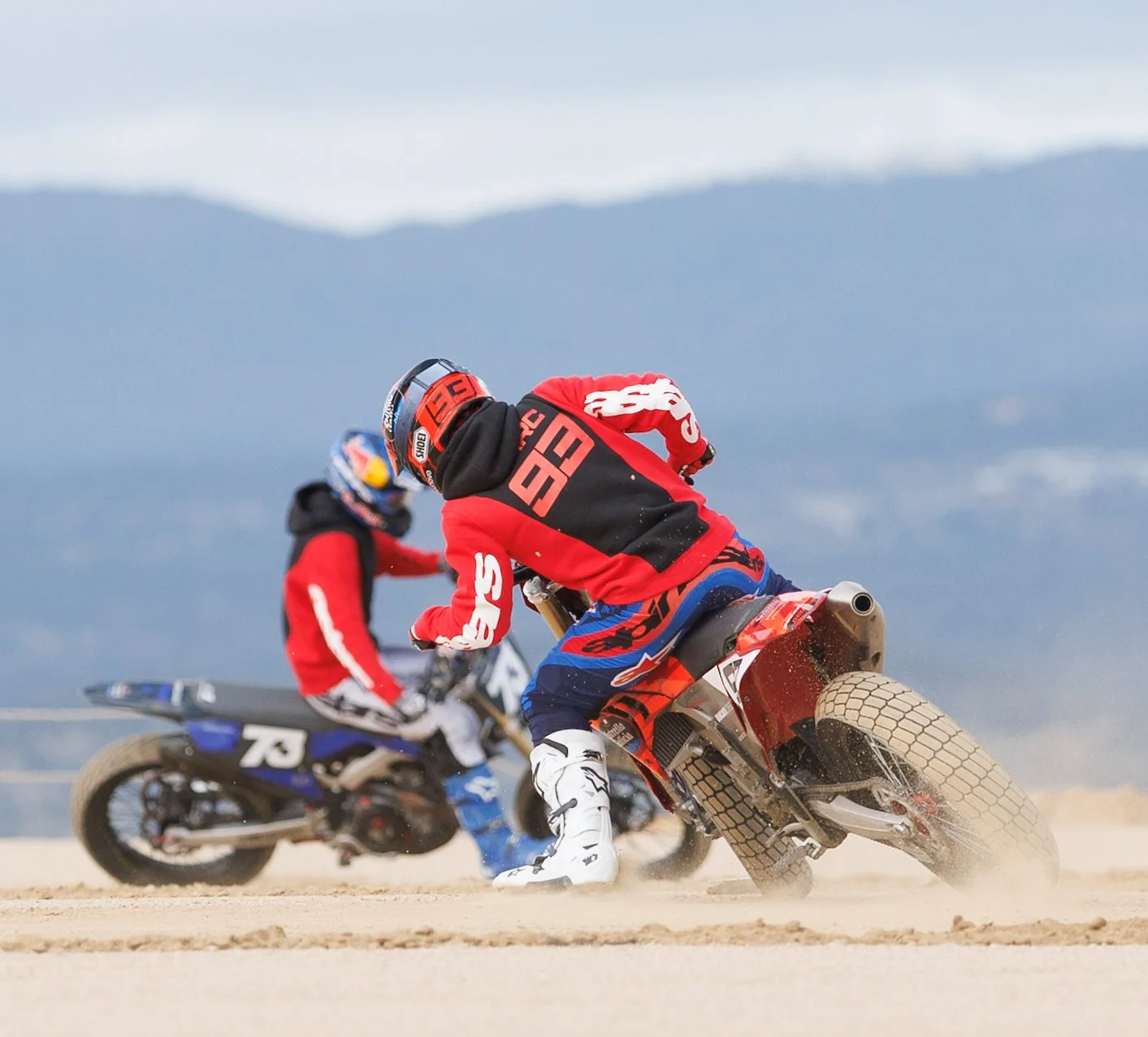 Dos motociclistas en una carrera en terreno arenoso con montañas al fondo.