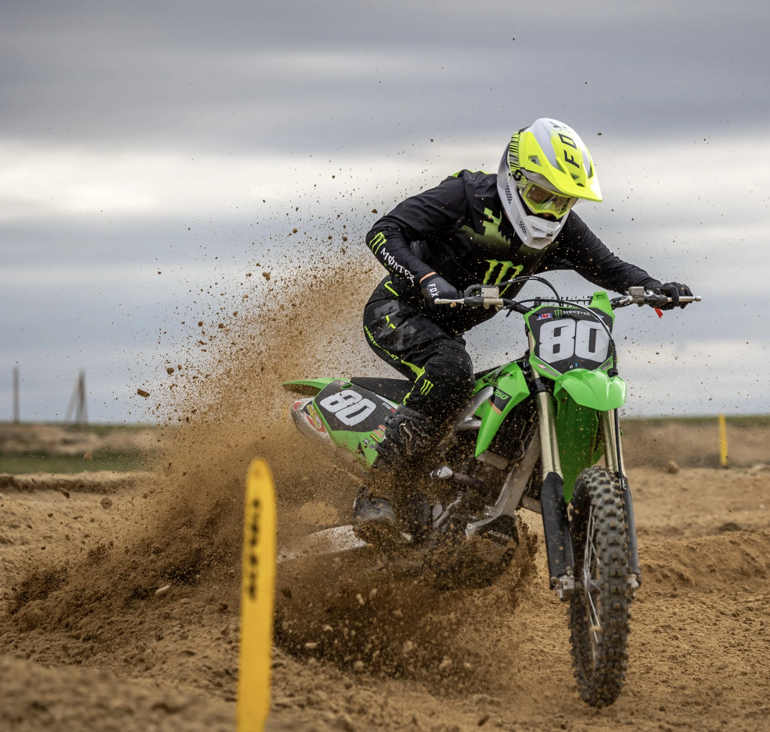 Piloto de motocross vestido con equipo negro y casco amarillo en una pista de tierra, montando una motocicleta verde con el número 80, en medio de una curva con polvo y arena levantados por la moto.