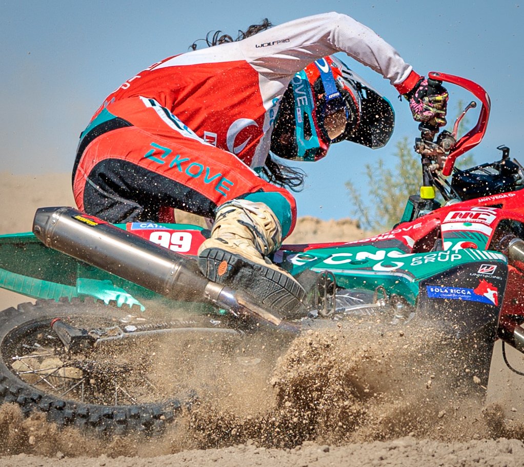 Un piloto de motocross en medio de una arena seca y polvorienta, inclinado hacia adelante a toda velocidad, usando un casco, guantes y un traje de protección con colores rojo, azul y blanco, conduciendo una moto de competición con marcas y patrocinad