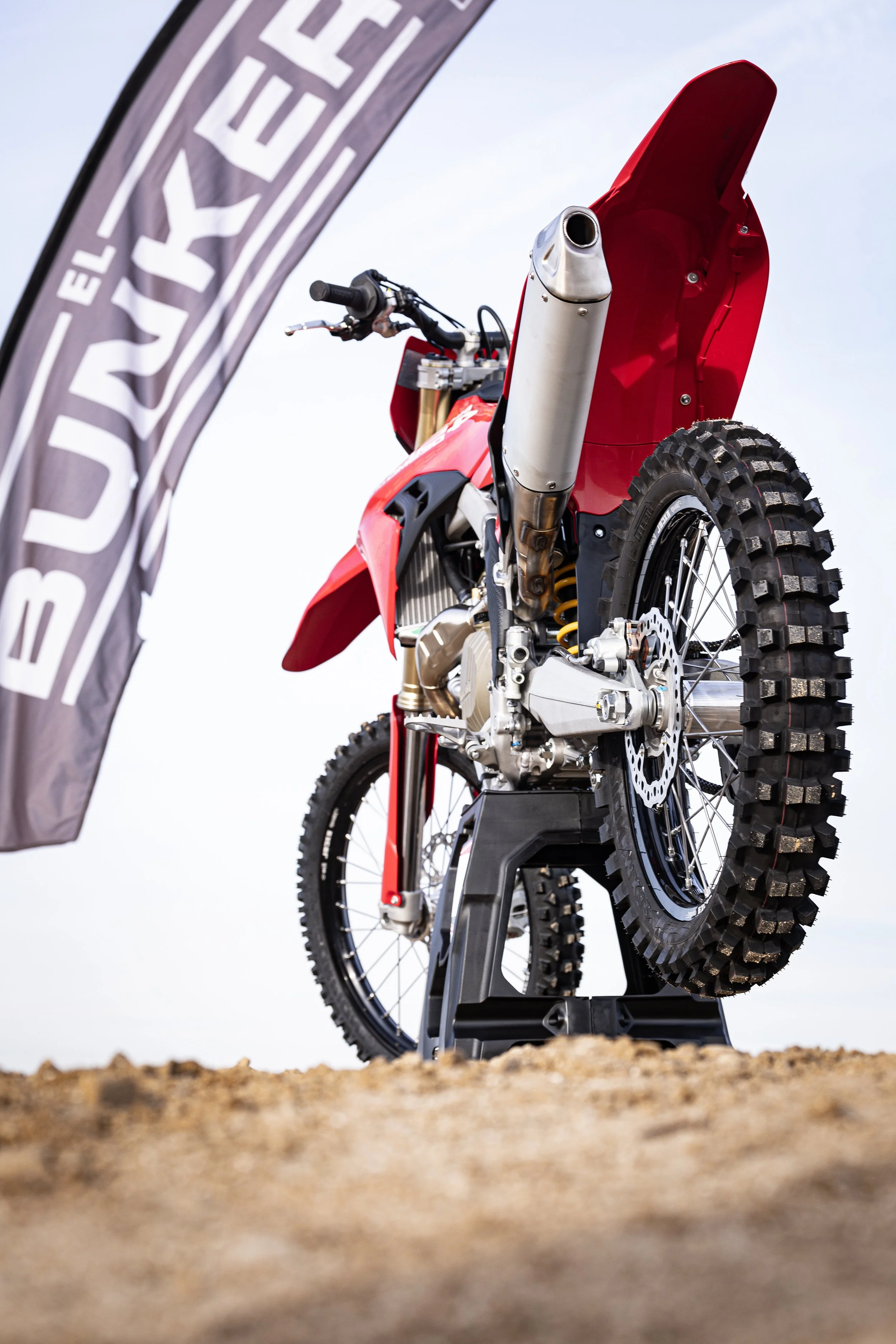 Motocicleta de motocross roja en exhibición, vista desde atrás, con bandera en el lado izquierdo que dice 'El JUEVES'.