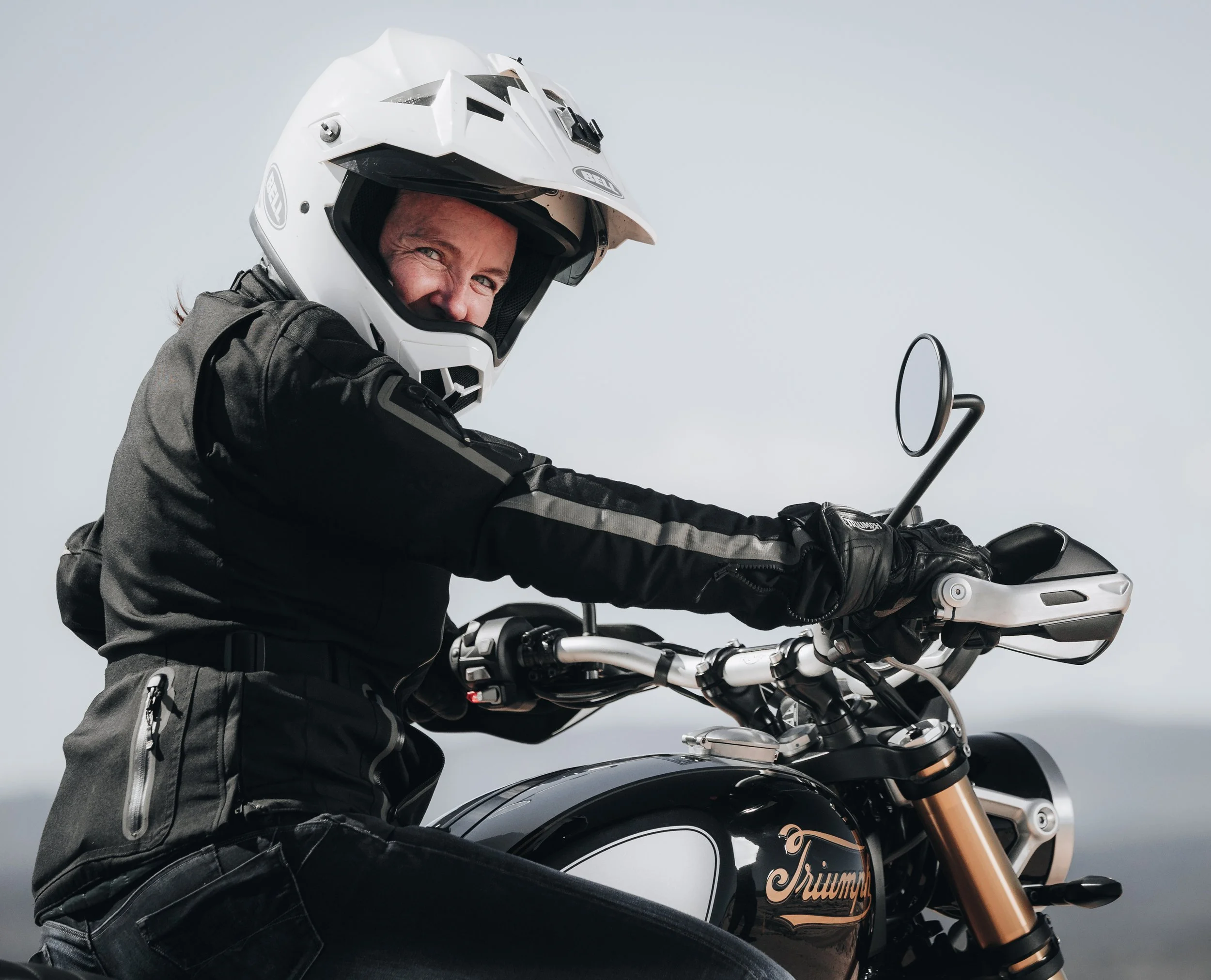 Una mujer con equipo de protección en una motocicleta Triumph, usando un casco y guantes, sonriendo mientras monta la moto.
