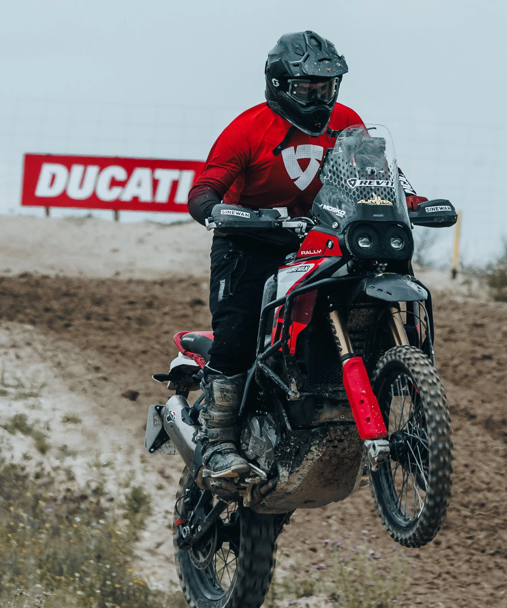 Persona montando una motocicleta de aventura en un terreno polvoriento, con casco y ropa de protección, en un evento patrocinado por Ducati.
