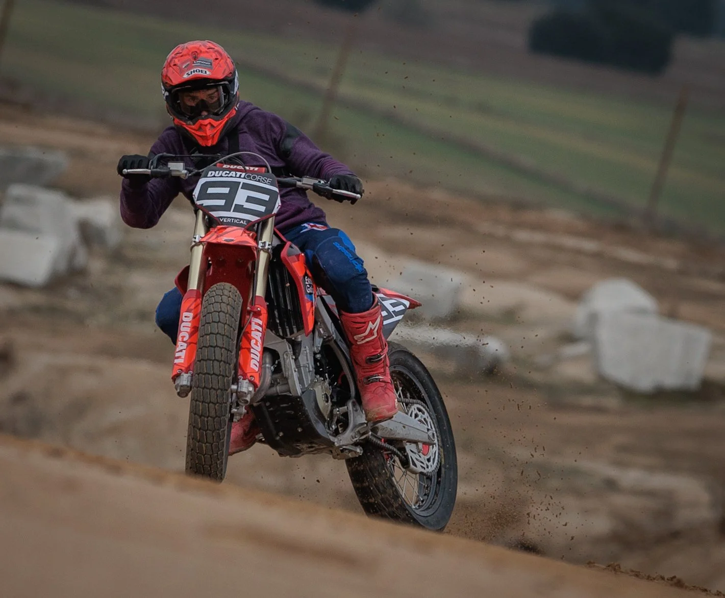 Piloto de motocicleta en carrera en un terreno de tierra, llevando casco rojo, chaqueta morada, pantalones azules, botas rojas y montando una motocicleta de competición naranja.