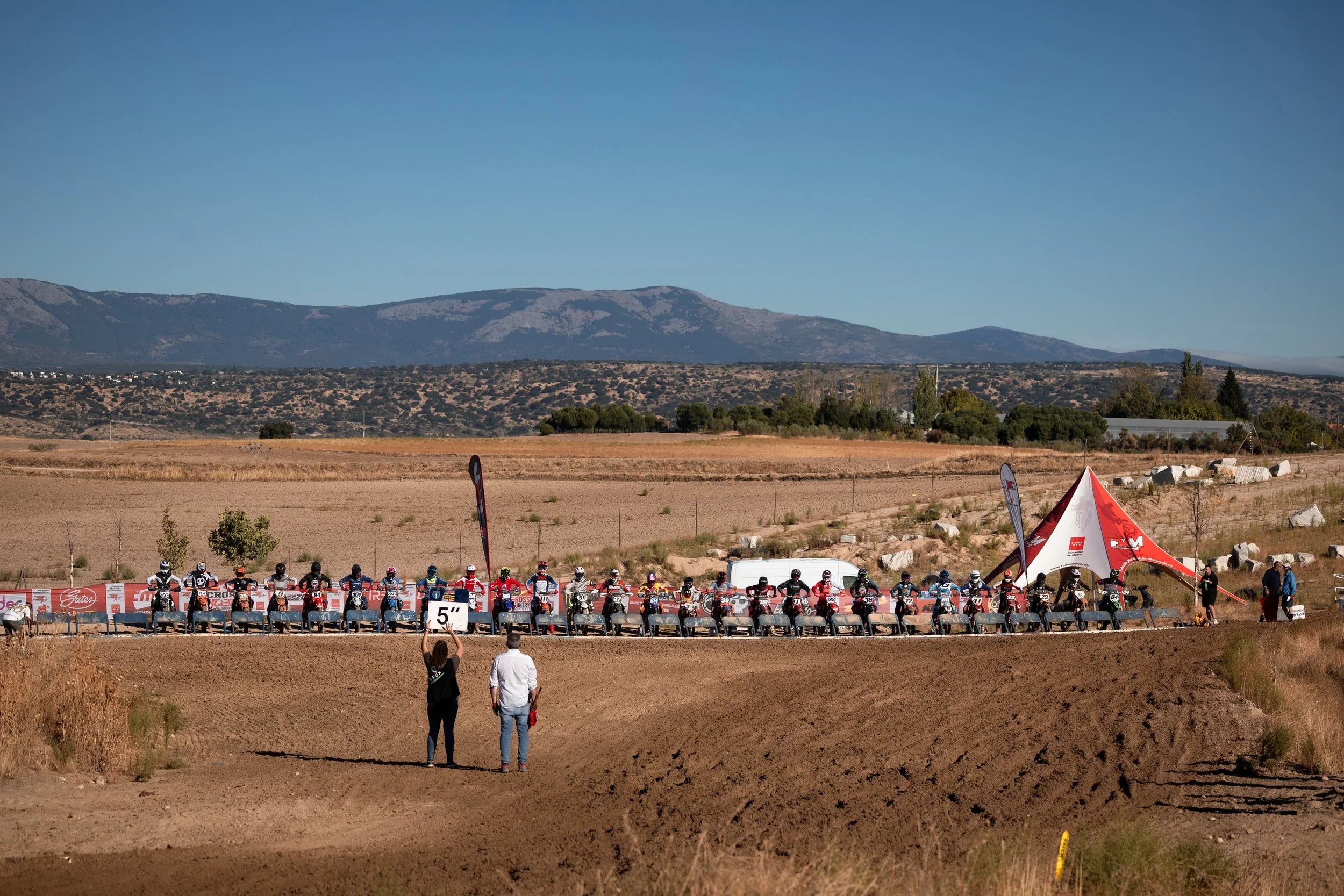 Inicio de una carrera de motocross en un terreno abierto con montañas al fondo, una fila de motociclistas en línea y un par de personas observando.