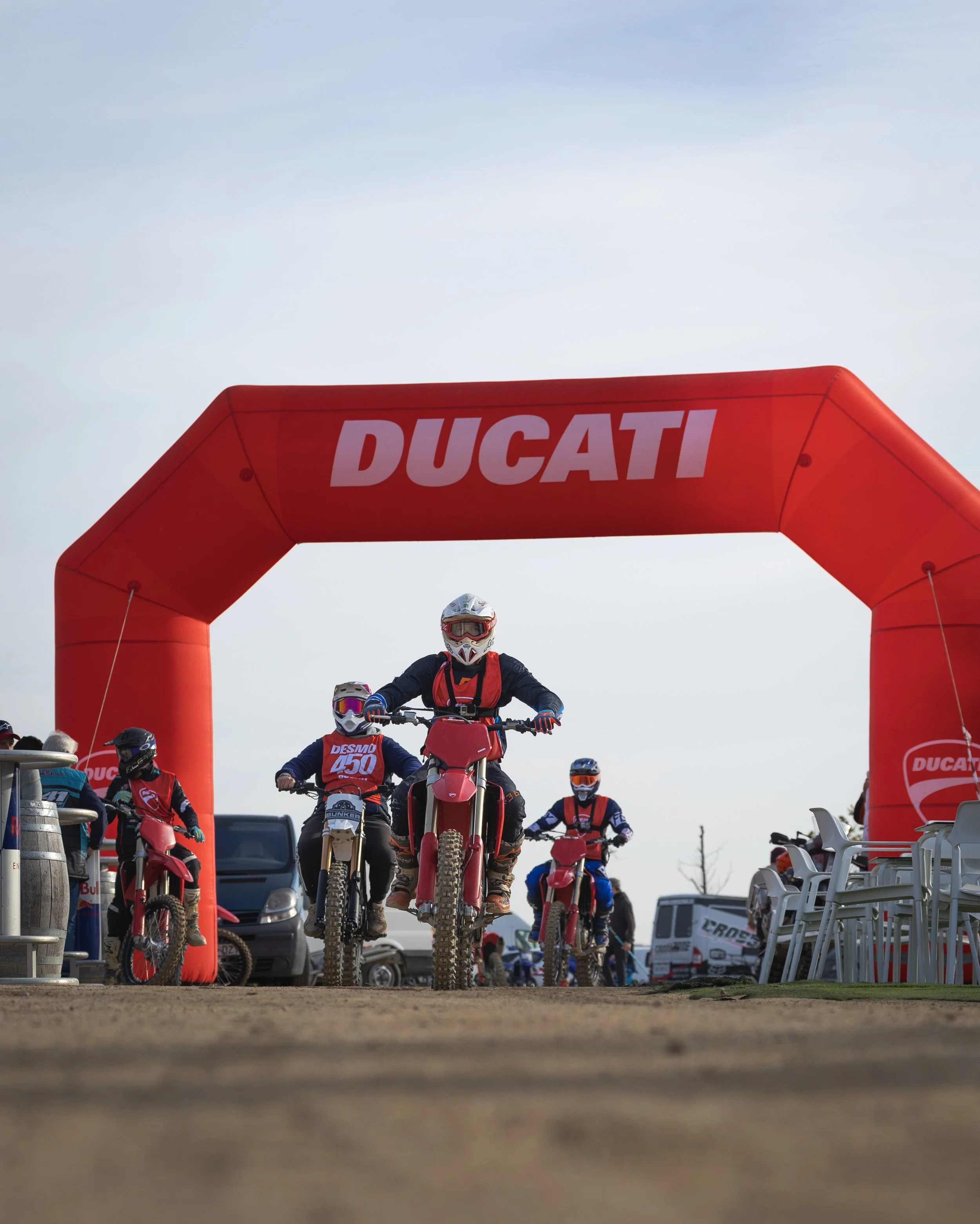 Carrera de motocicletas en una pista con un arco inflable rojo que lleva la marca 'DUCATI', varios motociclistas con casco y equipo de protección listos para comenzar la carrera, vehículos y sillas en el fondo.