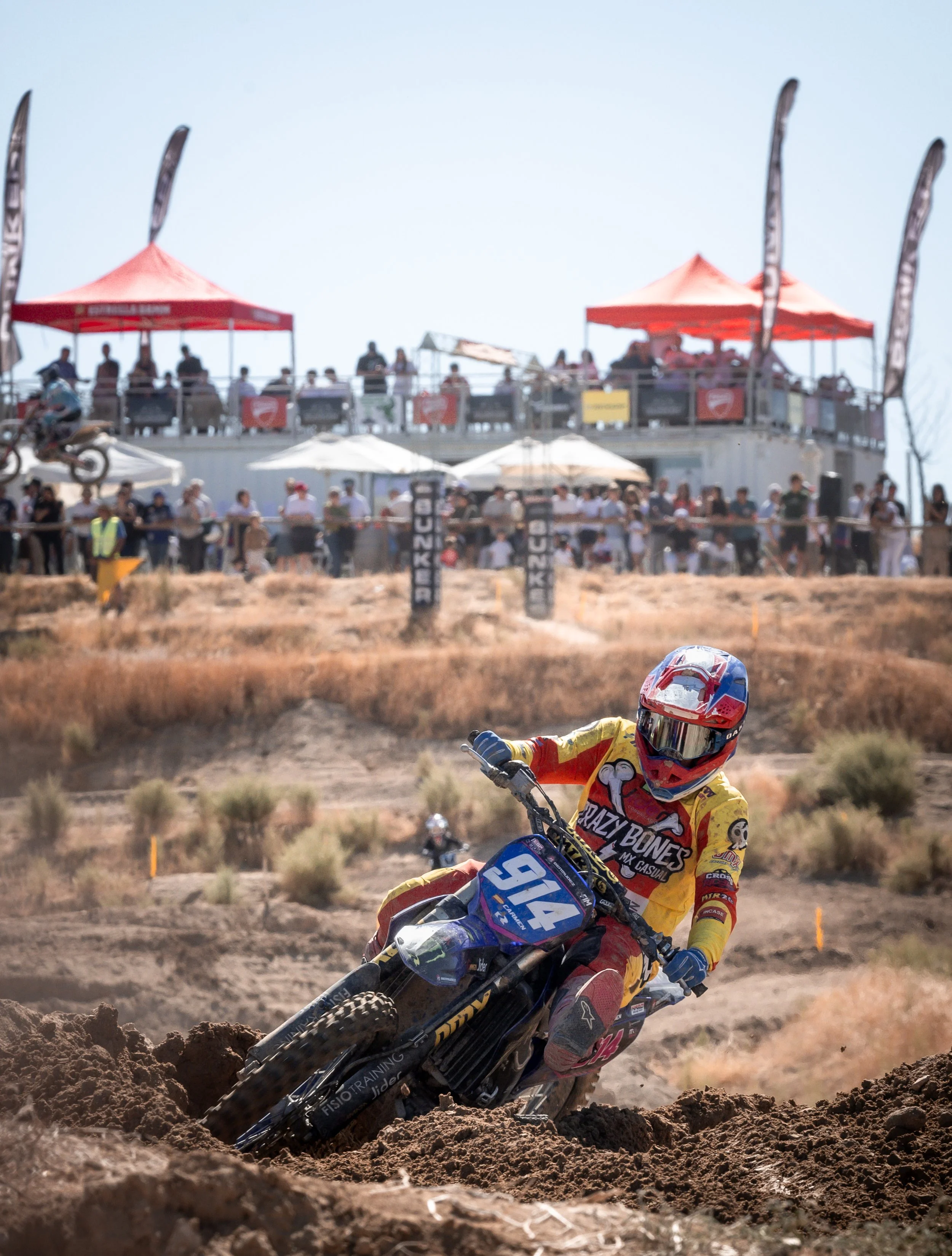 Carrera de motocross en un terreno arenoso con un corredor en una moto azul y roja, con el número 914, con espectadores en una tribuna larga y varias tiendas con toldos rojos y blancos en el fondo.