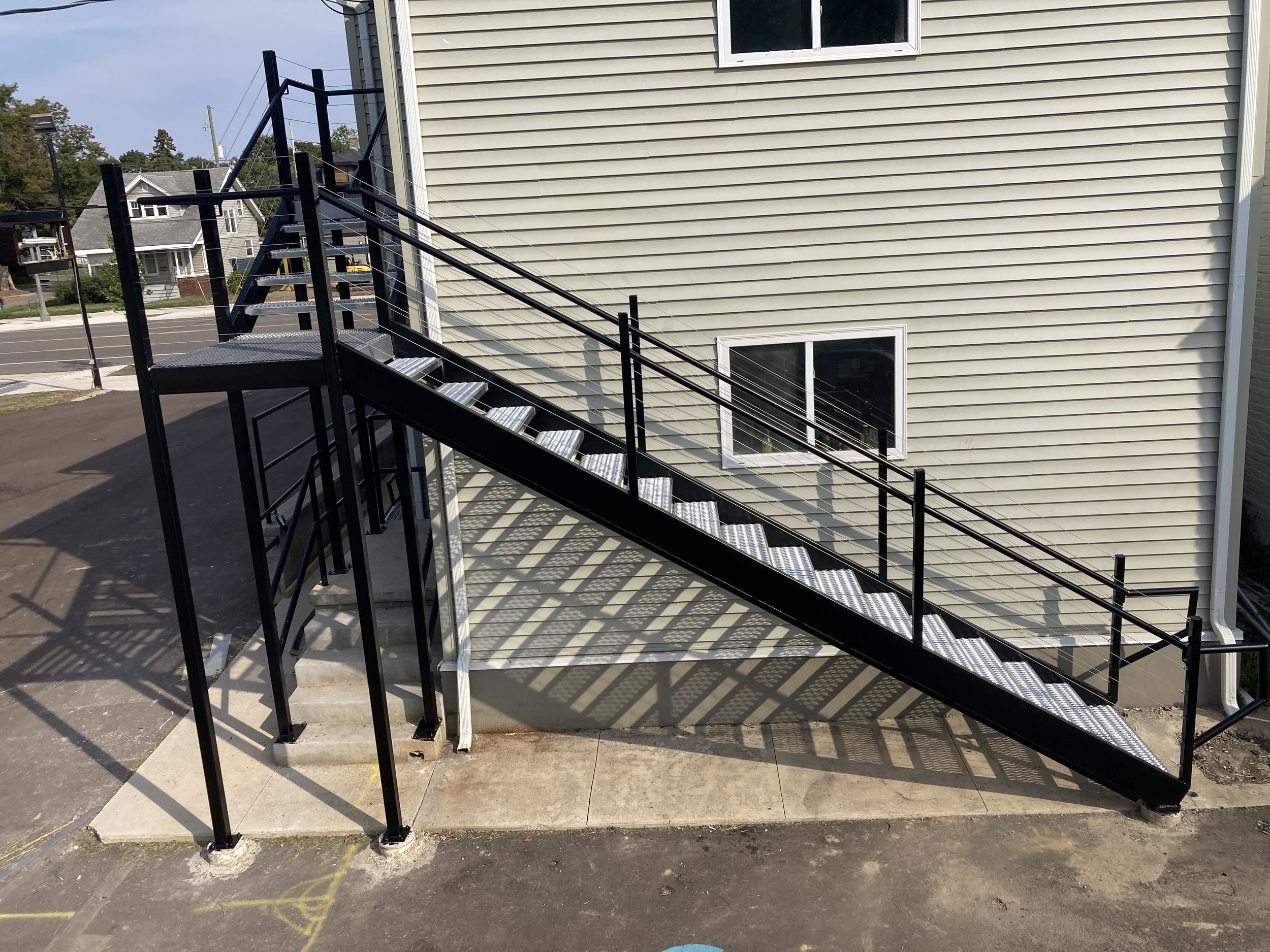 Black metal staircase with guardrails attached to the side of a beige house, leading from the ground level to an upper entrance.