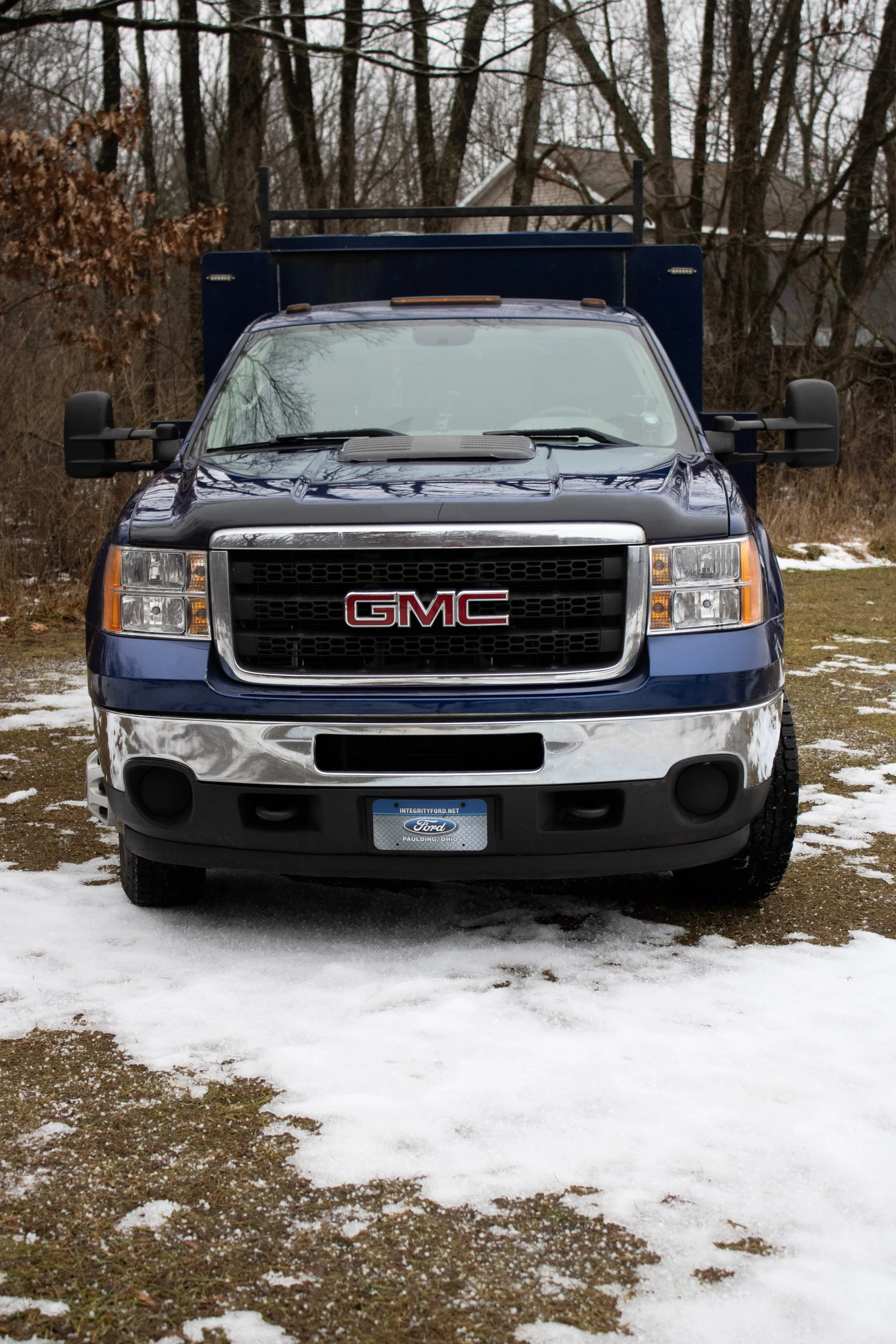 Front view of a dark blue GMC pickup truck parked on partially snow-covered ground, with leafless trees and a house in the background.