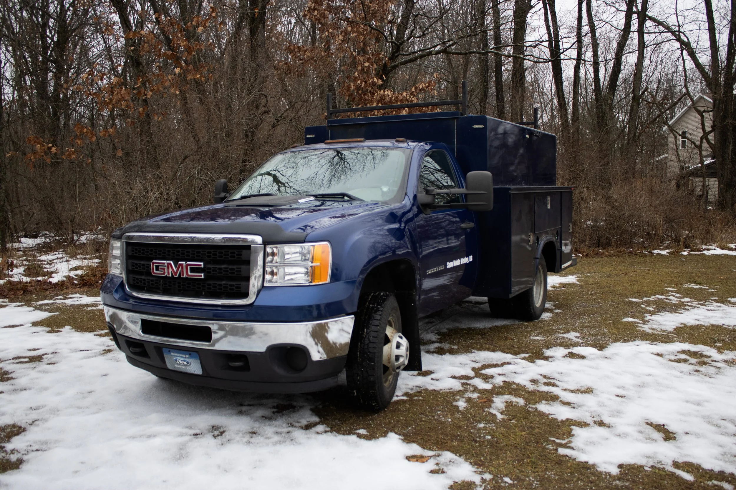 A blue GMC utility truck parked on a patchy snow-covered grassy area, with leafless trees and a house in the background.
