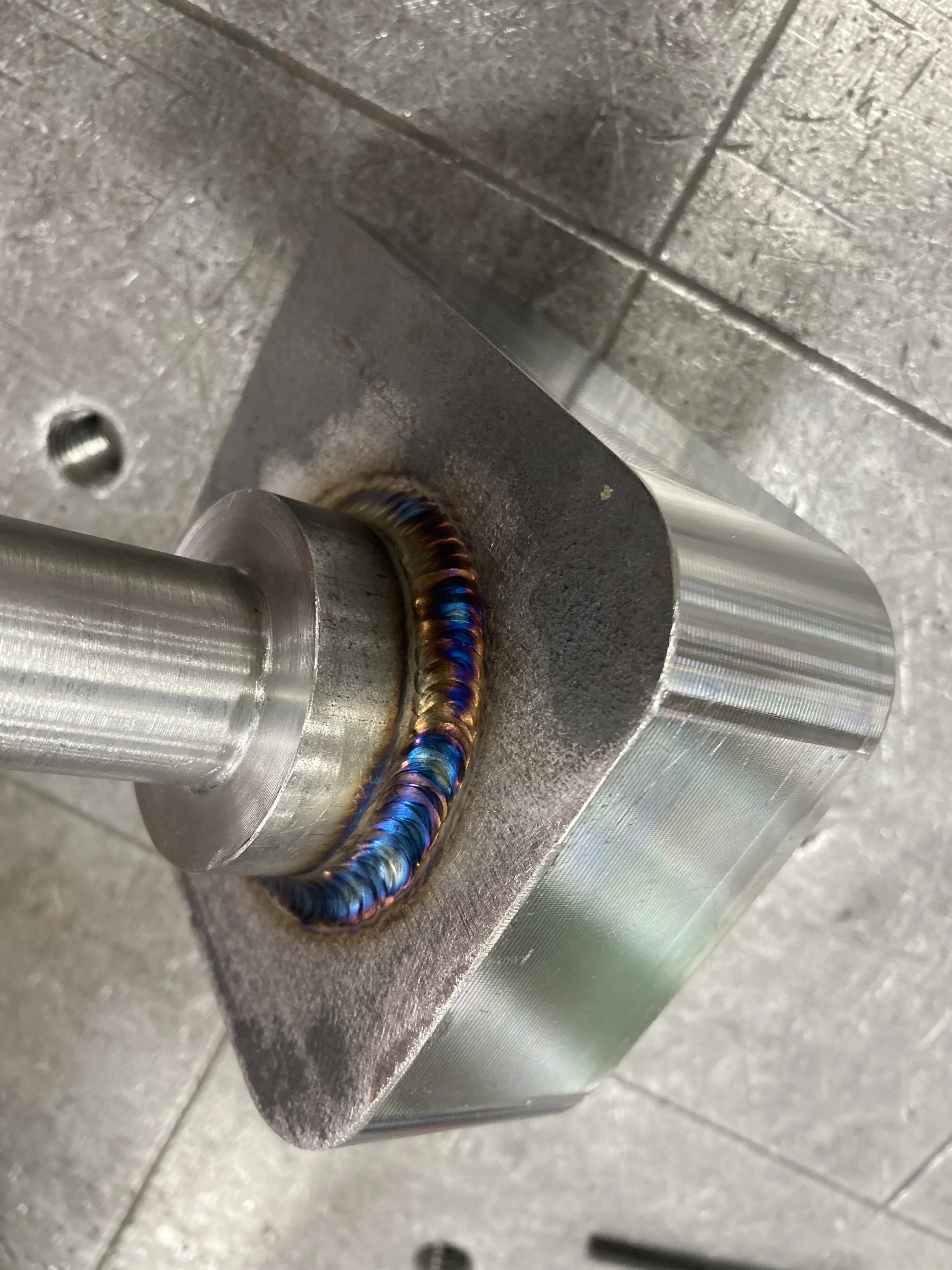 Close-up of a metal shaft welded to a metal block, showing colorful weld beads around the joint.