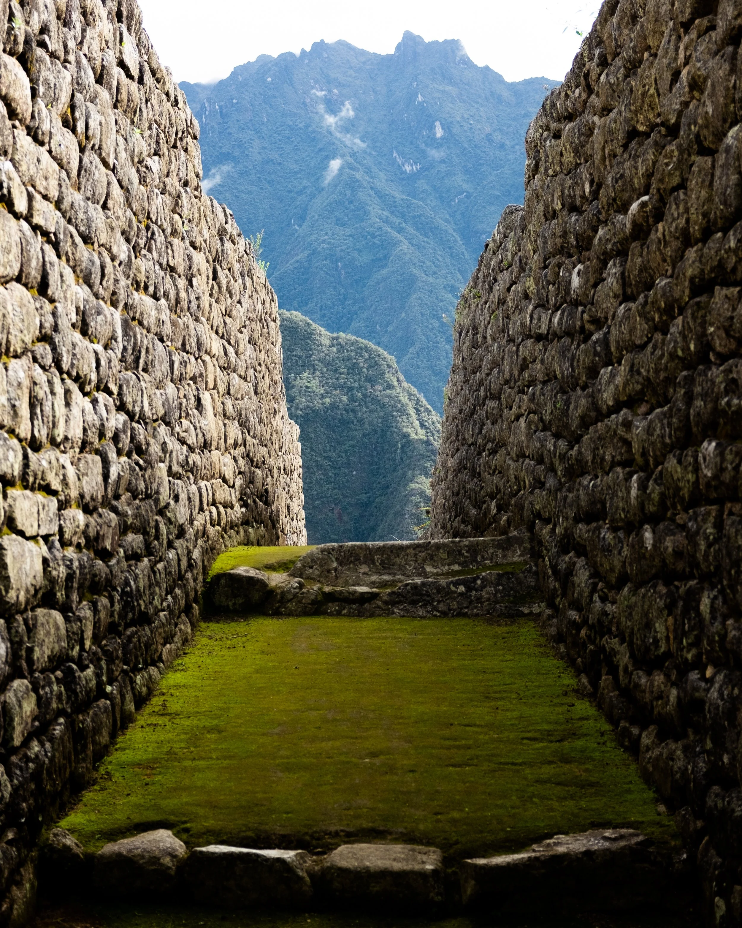 Ancient stone structures with moss-covered ground and towering mountains in the background.