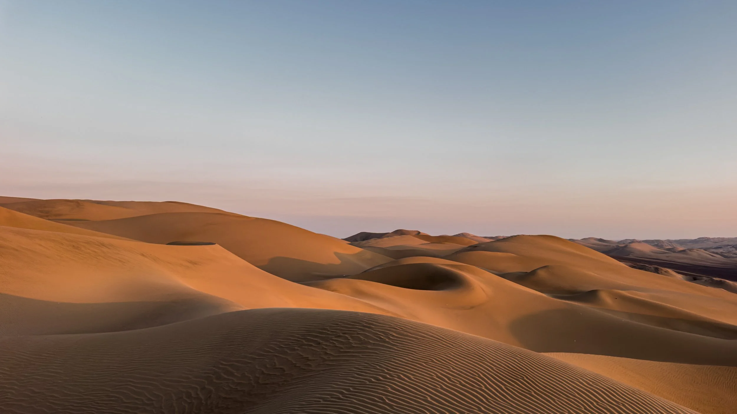 A vast desert with rolling sand dunes under a clear sky at sunset.