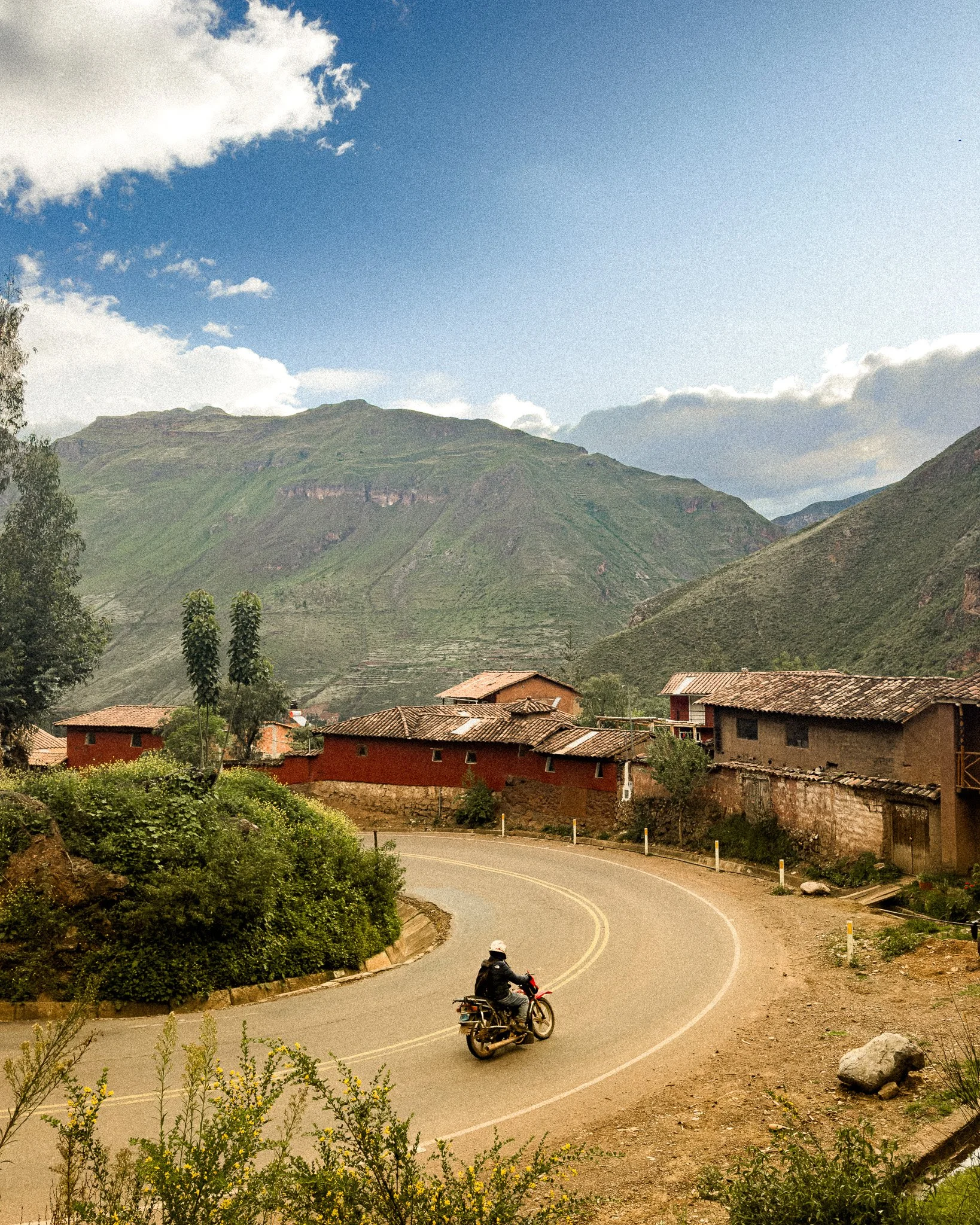 A person riding a motorcycle around a winding mountain road with green mountains and a partly cloudy sky in the background.