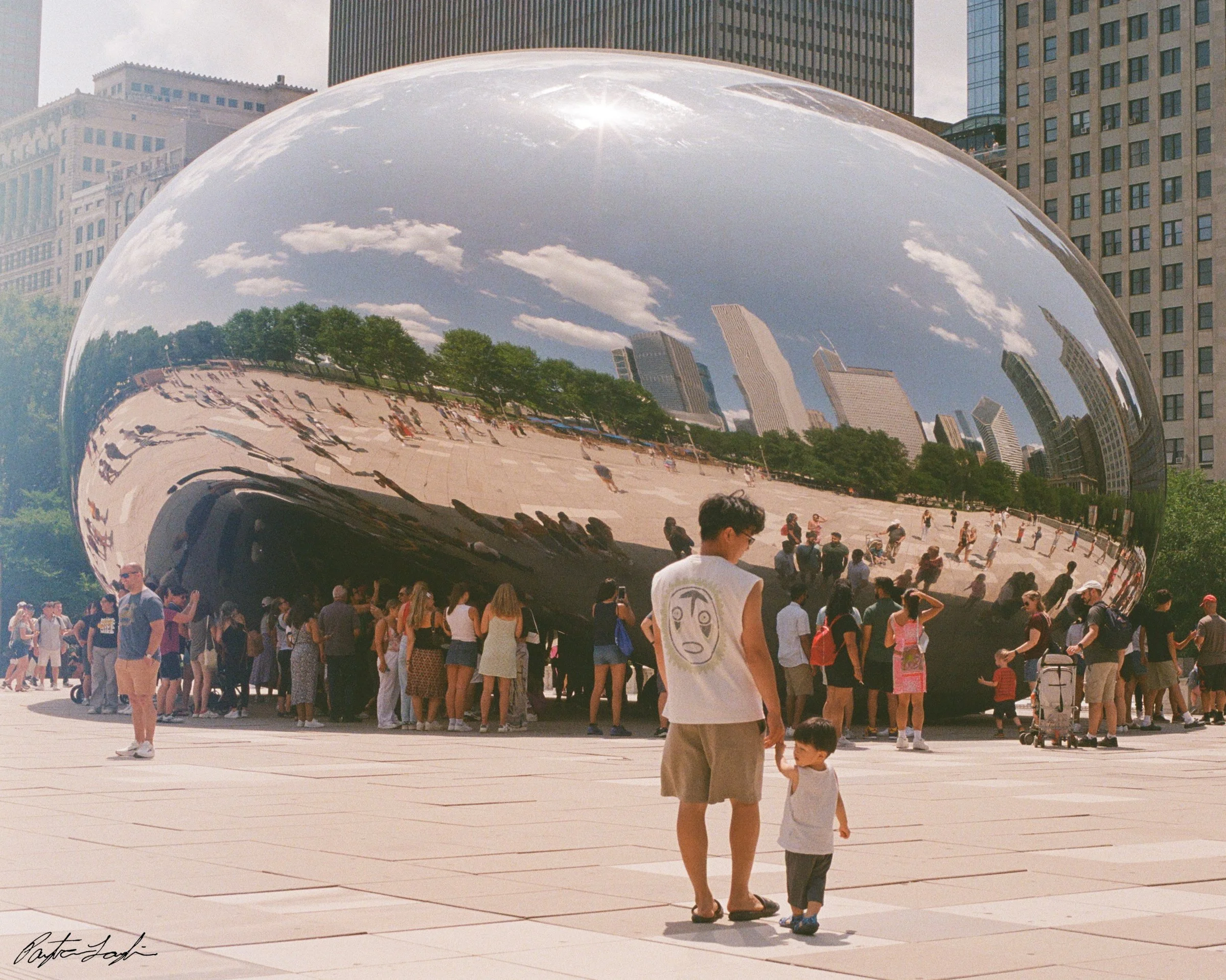 A large reflective sculpture in an urban plaza with people gathered around, reflecting the cityscape and sky above.