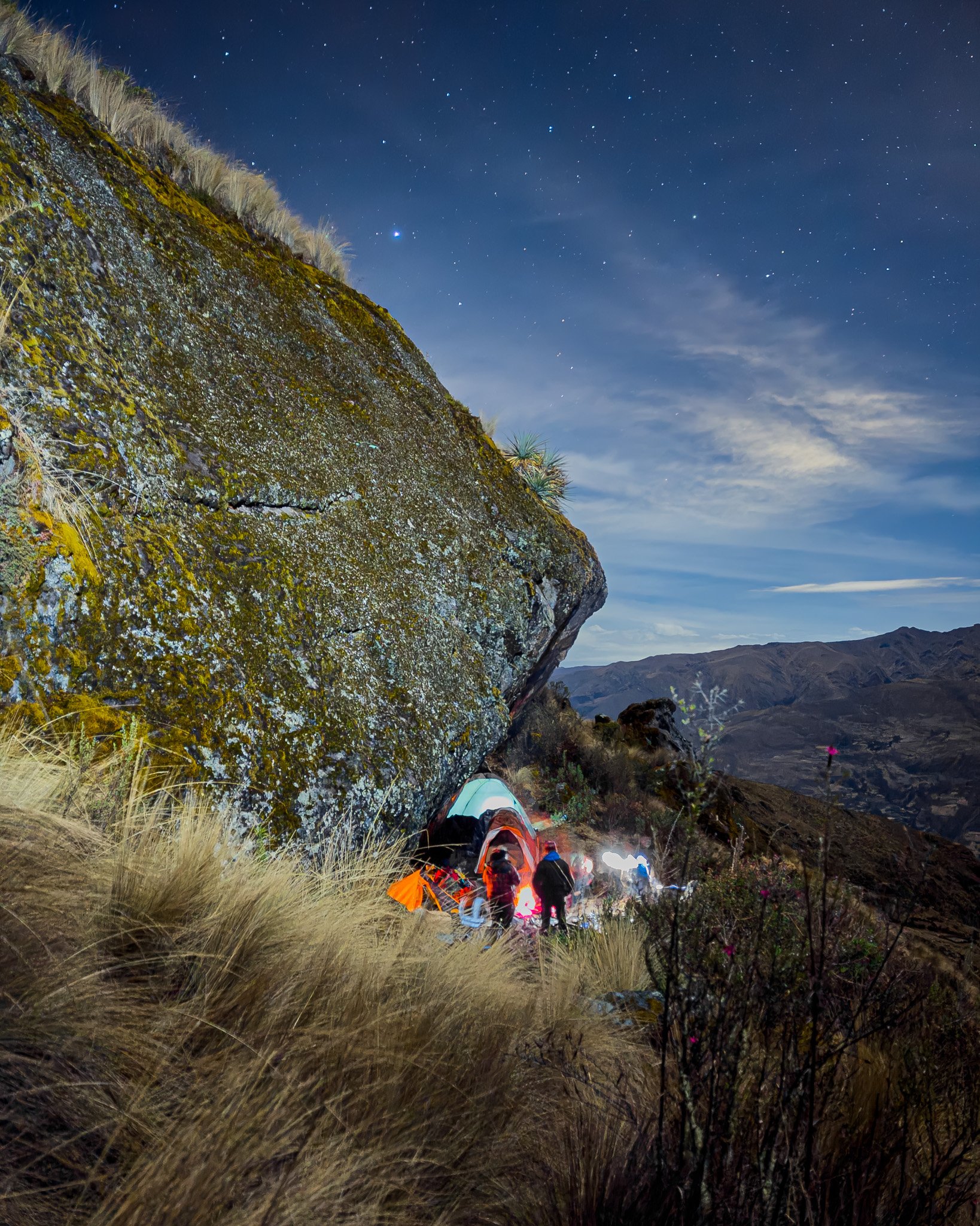 Camping tents set up on a hillside at night under a starry sky, with large moss-covered rock and dry grass in the foreground.