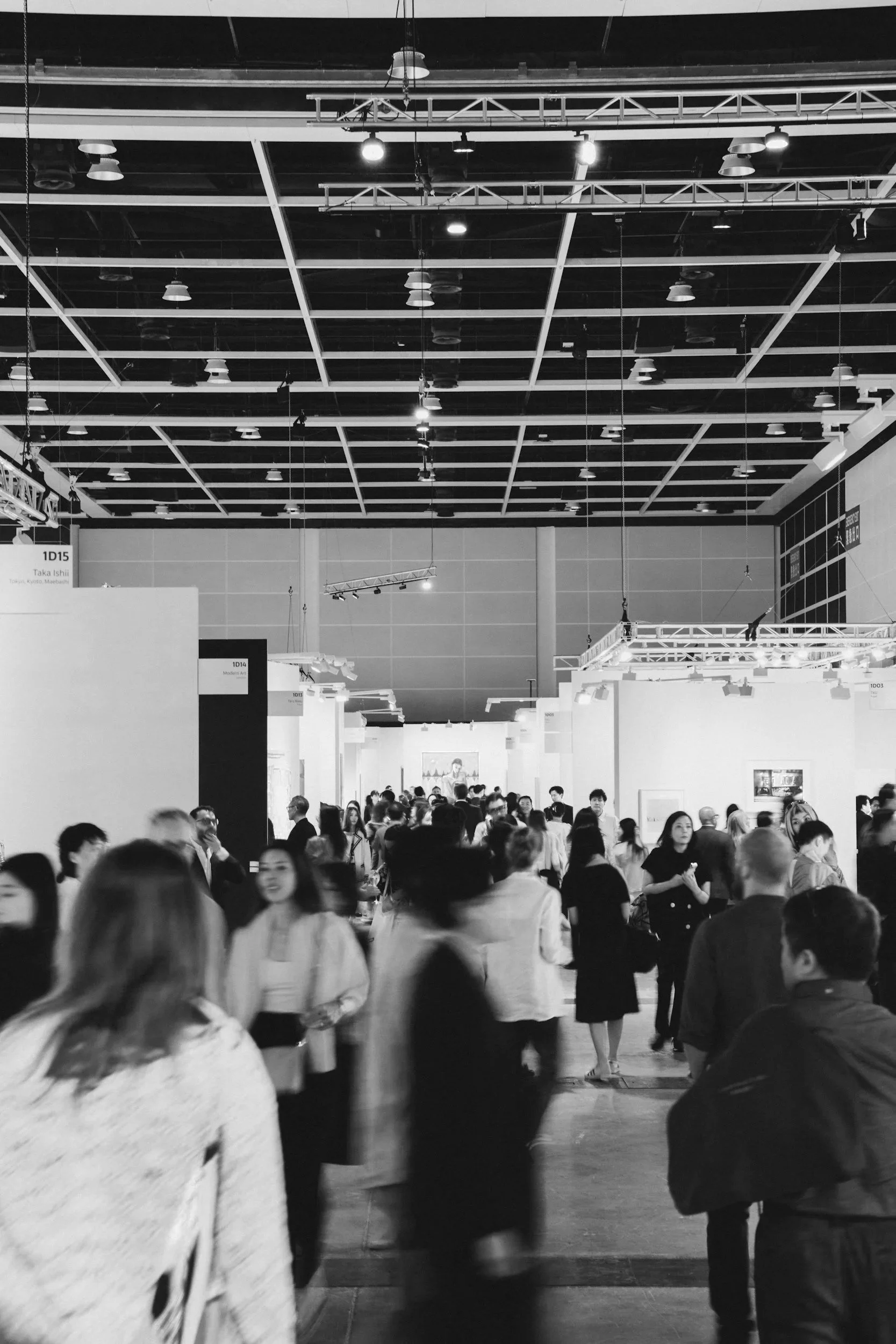 A crowded exhibition hall with many people walking and socializing, white display booths, and a high ceiling with exposed beams and lighting.