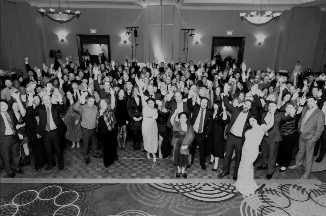 Large group of people gathered in a banquet hall, celebrating and waving at the camera.