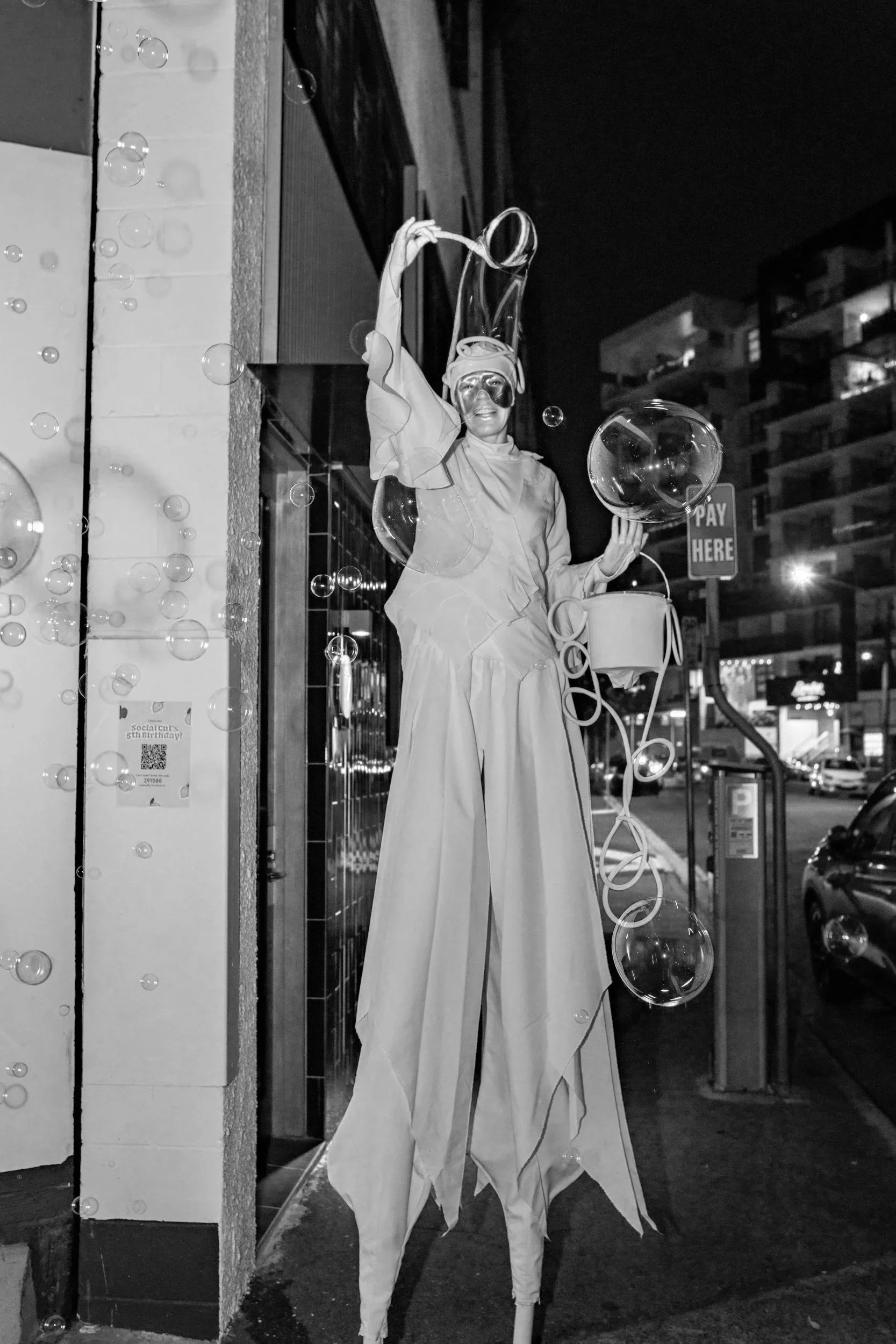 A street performer on stilts dressed as an abstract artistic figure holding a bubble wand and transparent balloons, illuminated at night with buildings and cars in the background.