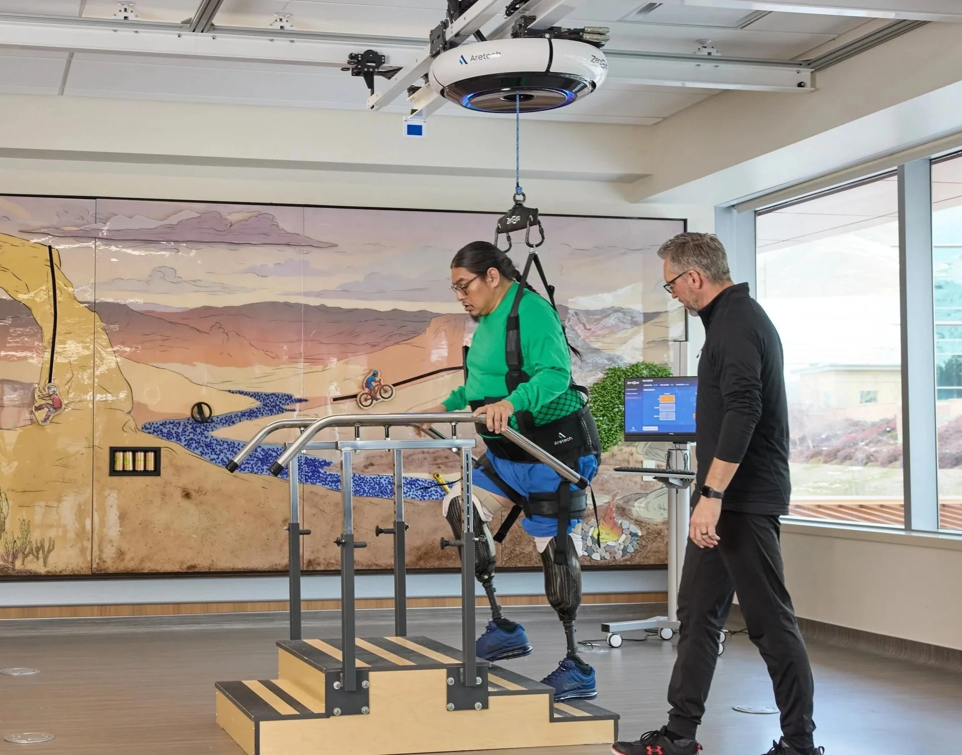 A person with a prosthetic leg practicing walking on a treadmill during a clinical trial. An engineer or researcher observes nearby. They are inside a modern room with large windows and a mural of mountains and outdoor scenes on the wall.