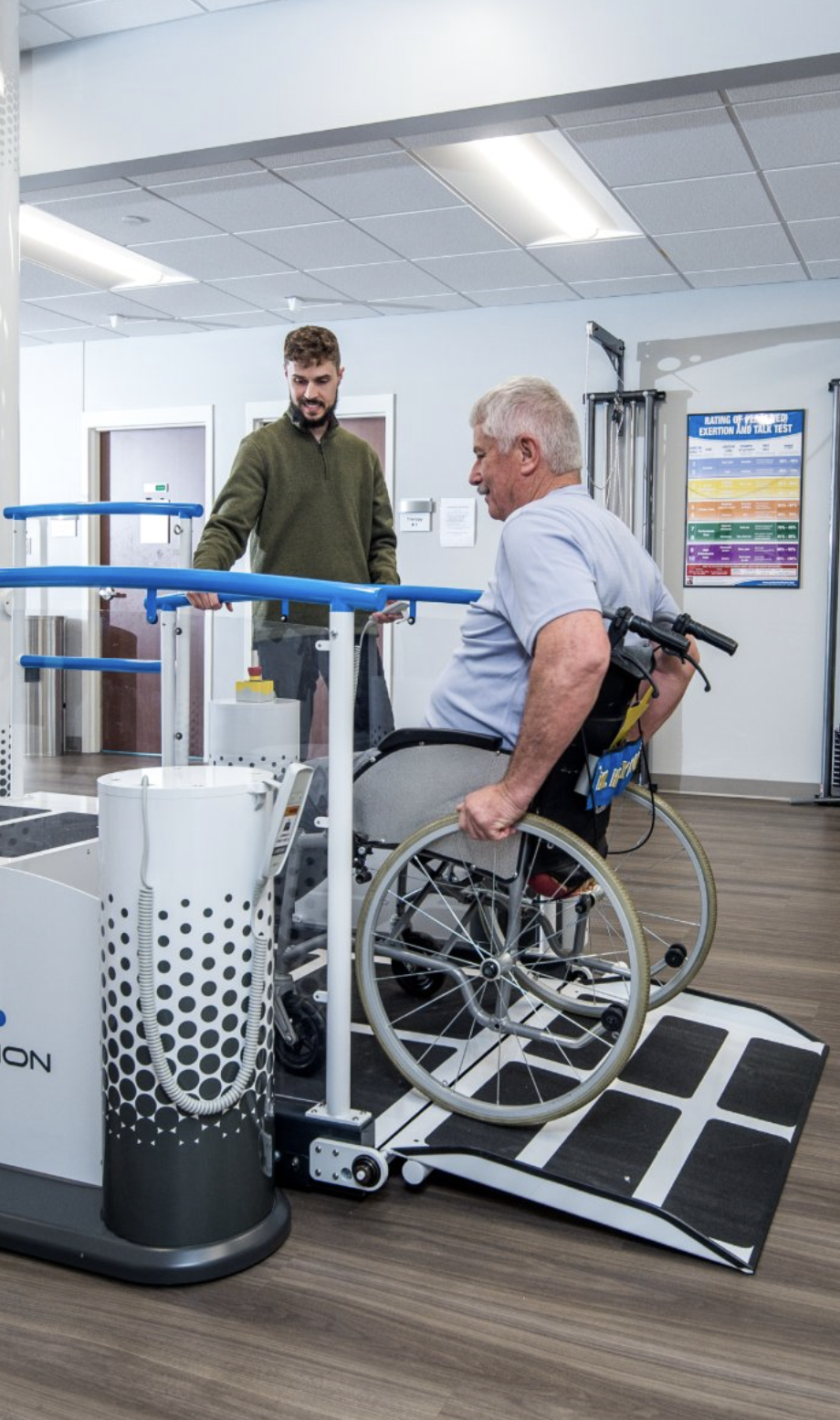 A man in a wheelchair using an accessible hand strength test machine in a medical or therapy facility, with a staff member assisting him.