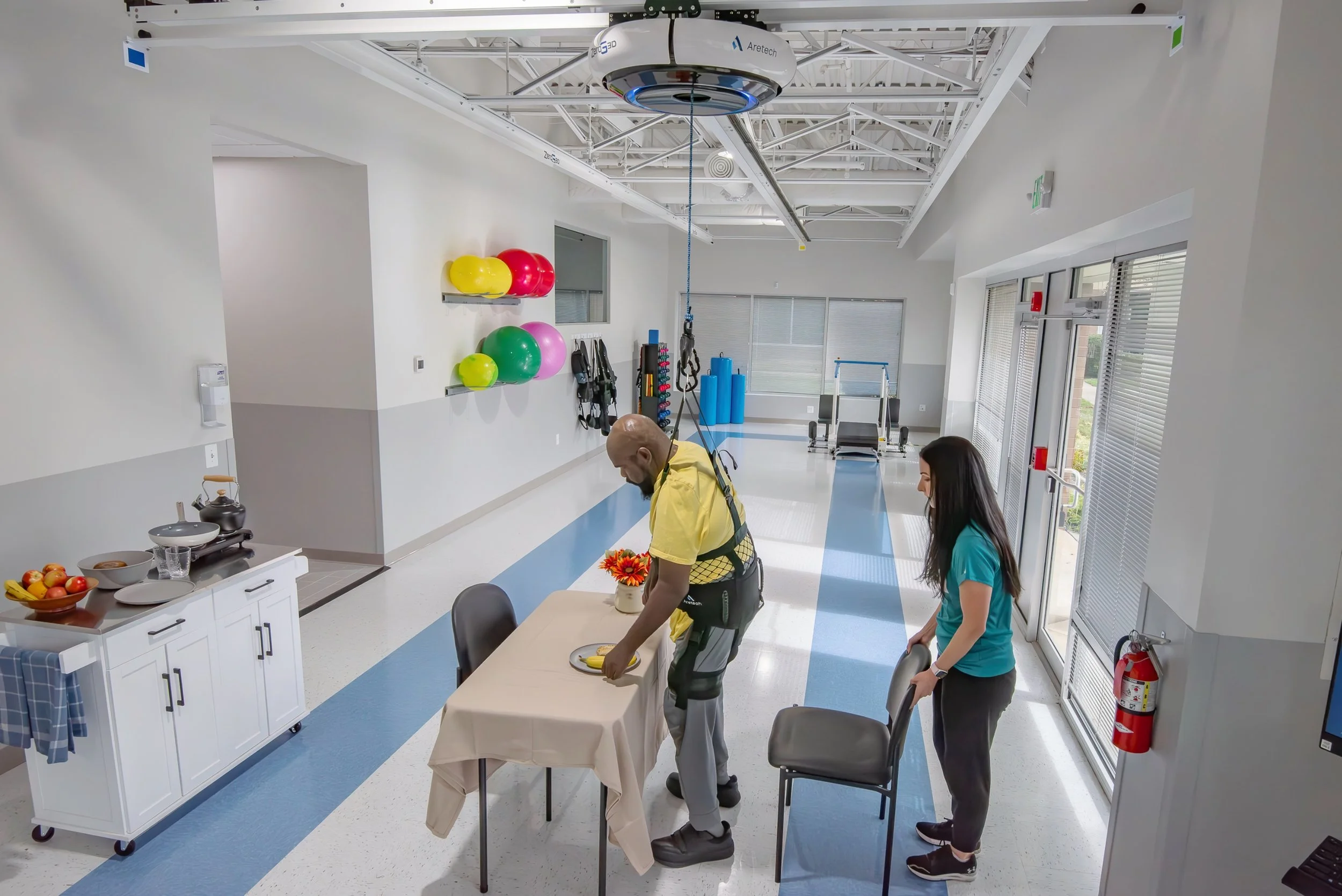 A therapy or rehab room with two people, a man and a woman, at a table with a tan tablecloth. The man is in a wheelchair and is reaching for a banana on a plate. The woman is observing, and another person is walking past. The room has white walls, shelves with colorful therapy balls, and sports equipment. Large windows with blinds and a door with glass panels are visible.