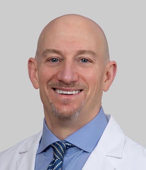 A smiling male healthcare professional with a bald head, light blue collared shirt, striped tie, and white coat, against a gray background.
