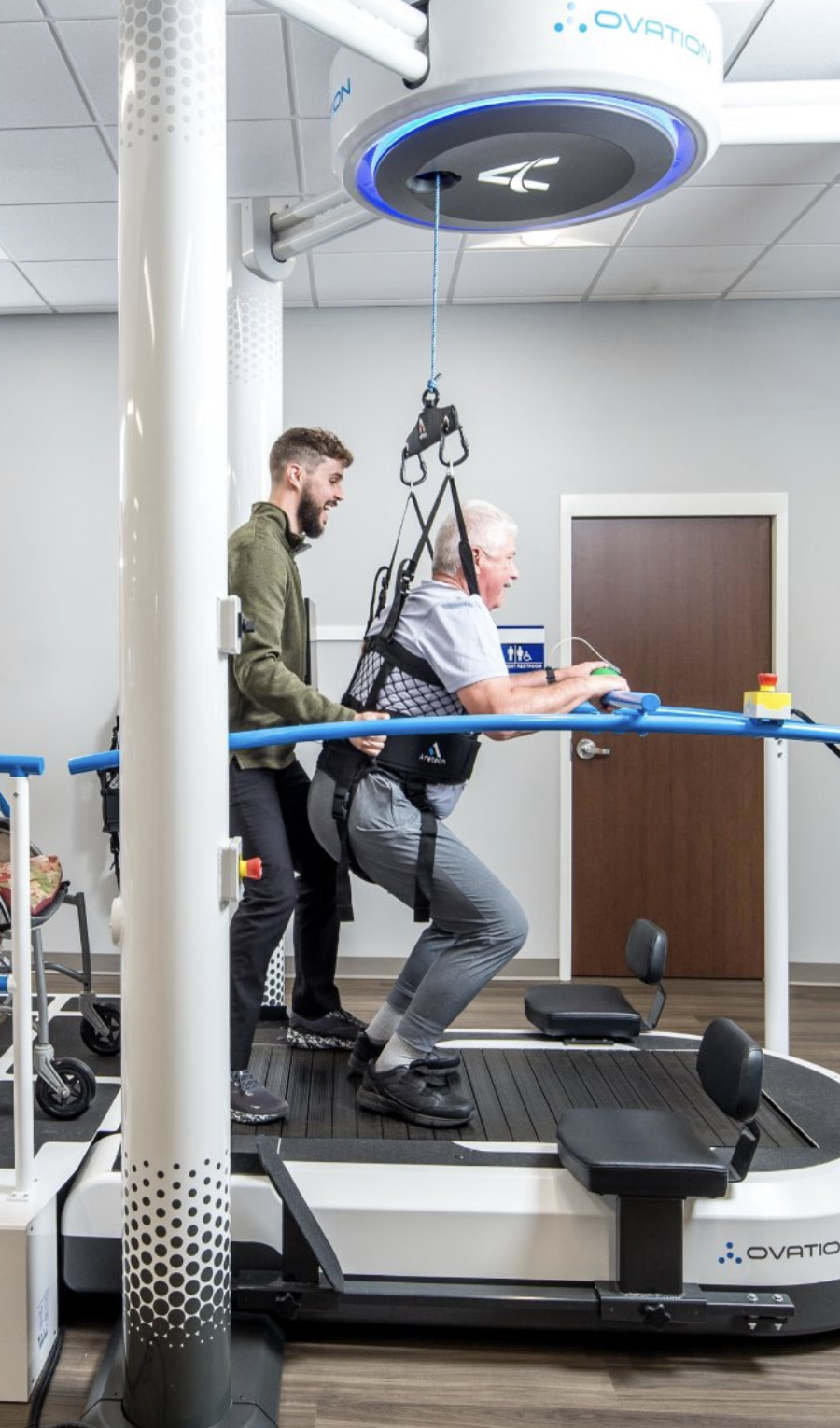 An elderly man is using a treadmill with assistance from a healthcare worker during a therapy session. The man is walking while connected to a harness system attached to a ceiling-mounted device, helping support his weight.