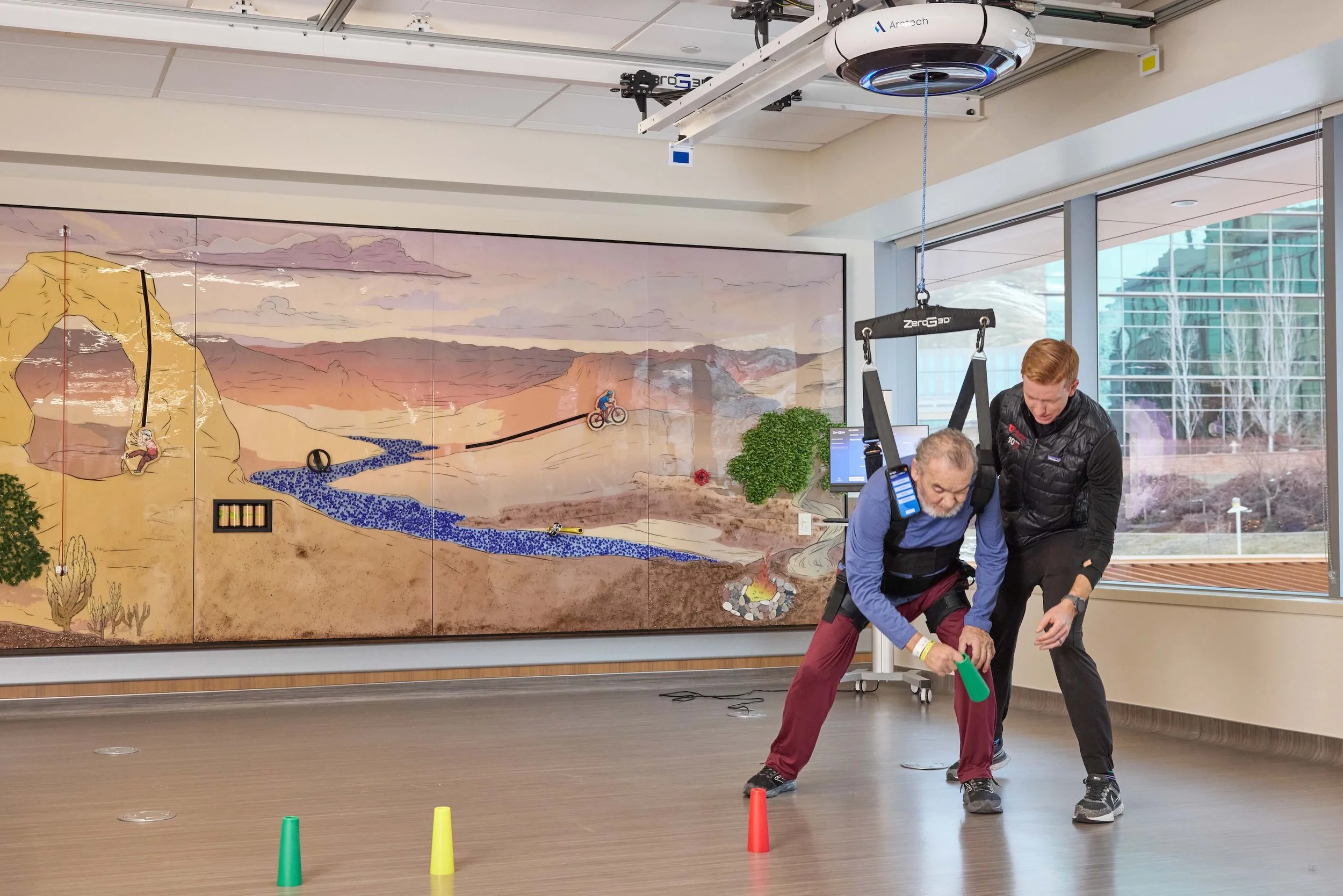 Two men in a room with a large mural of mountains and a river on the wall, engaging in a rock climbing or adventurous activity simulation with cones on the floor, a harness hanging from the ceiling, and a tablet or device nearby.