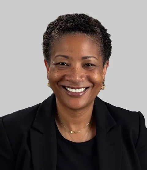 A woman with short, curly black hair and a bright smile, wearing a black blazer and gold jewelry, against a plain gray background.