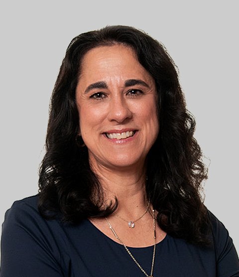A woman with long dark hair smiling, wearing a dark blue top and a necklace, against a plain gray background.