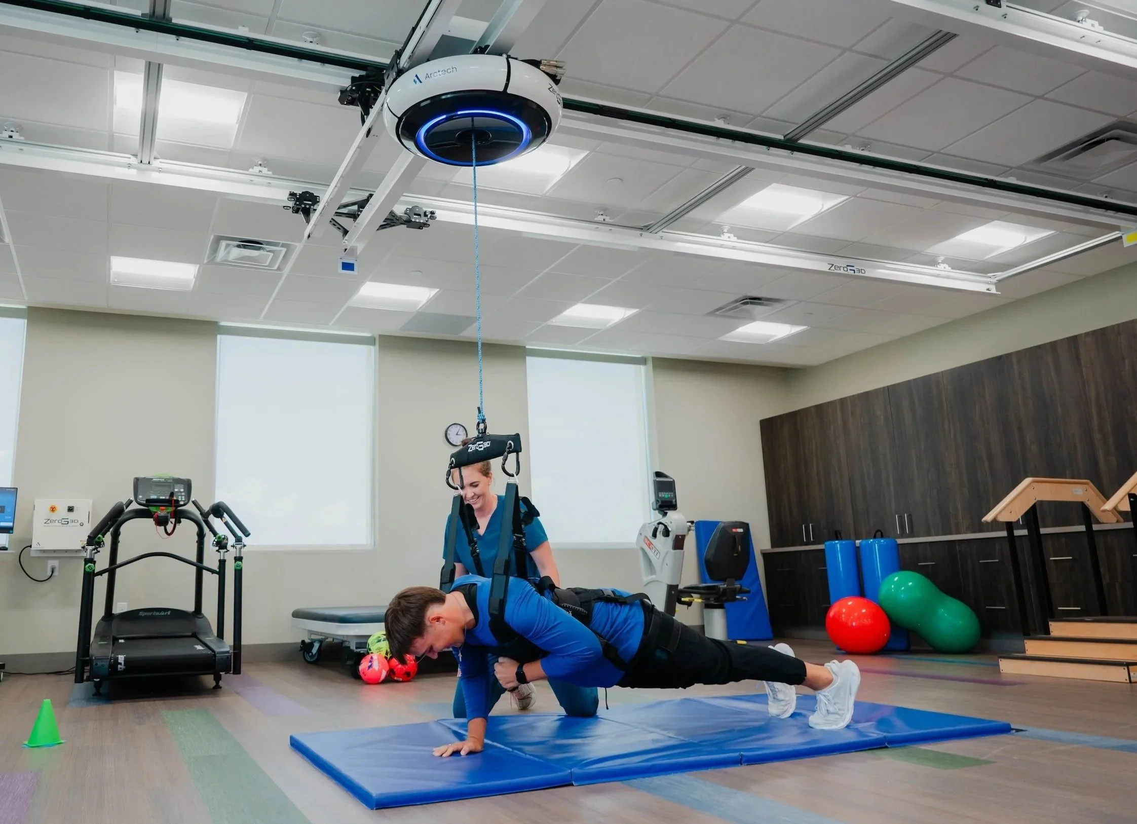 A woman and a boy are practicing a plank exercise in a physical therapy or rehabilitation room. The woman is on her hands and toes on a blue mat, smiling. The boy is in front of her, also on the mat. Medical and exercise equipment, including a treadmill, stability balls, and a harness, are visible in the room.