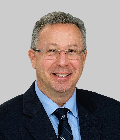 A middle-aged man with glasses, wearing a suit, light blue shirt, and a dark striped tie, smiling in front of a gray background.