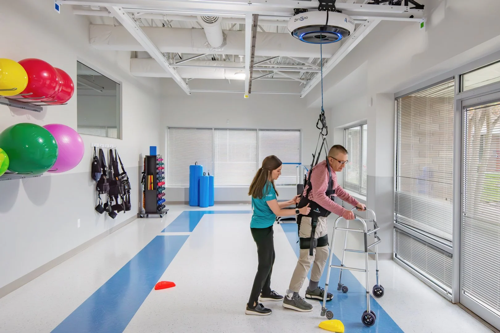 A man walking with a walker assisted by a female physical therapist in a rehab center, with therapy equipment and exercise balls in the background.