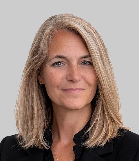 Headshot of a woman with blonde hair wearing a black blazer, smiling lightly, against a gray background.