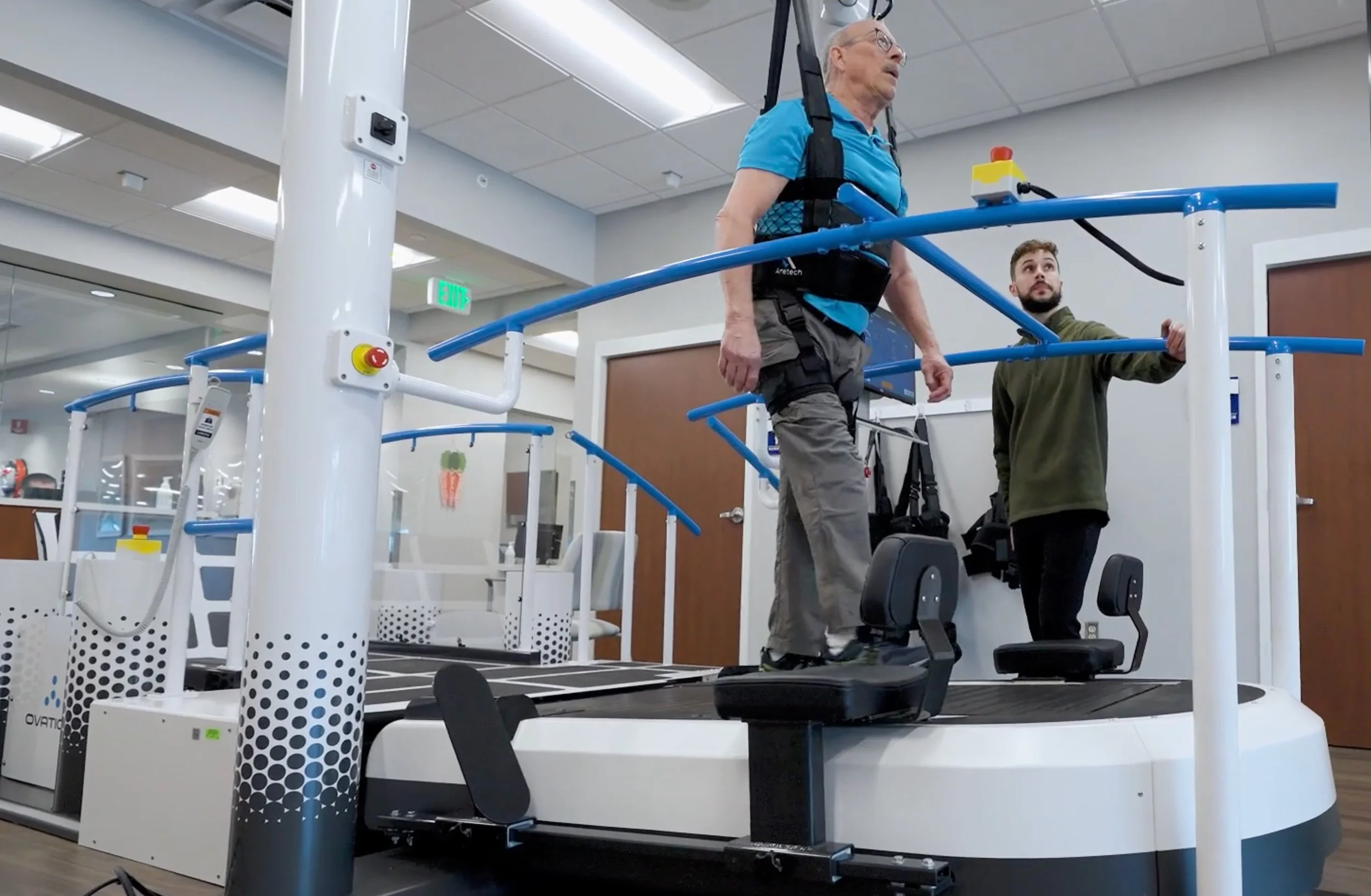 An elderly man wearing a blue shirt and gray shorts walking on a treadmill with a harness, supervised by a young man in a green jacket in a medical or rehabilitation facility.