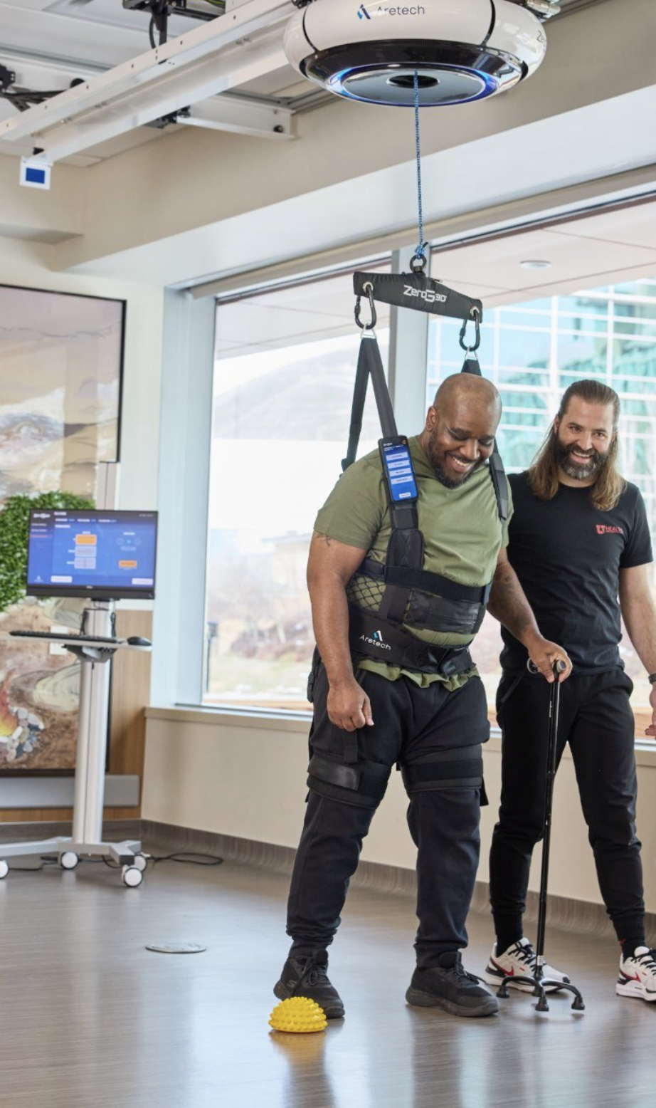 A man in a green shirt and black pants is participating in a physical therapy session using a harness system suspended from the ceiling, with a therapist beside him. The man is smiling and balancing on a yellow textured dome, while the therapist watches and assists. There is a monitor on a rolling stand displaying a program or data in the background.