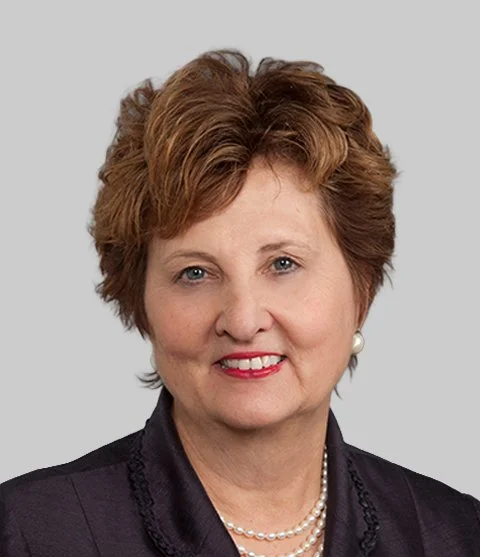 Portrait of an older woman with short, curly light brown hair, wearing a dark blazer, pearl earrings, and a pearl necklace, smiling against a plain light gray background.