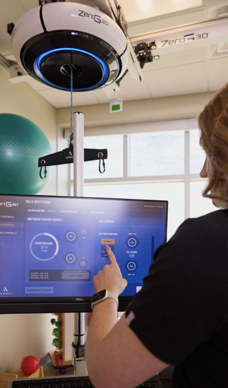 A person operating a computerized control panel for a medical or physical therapy device, with a robotic or automated system overhead marked 'zeroG3D,' and a green exercise ball in the background in a clinical or rehabilitation setting.