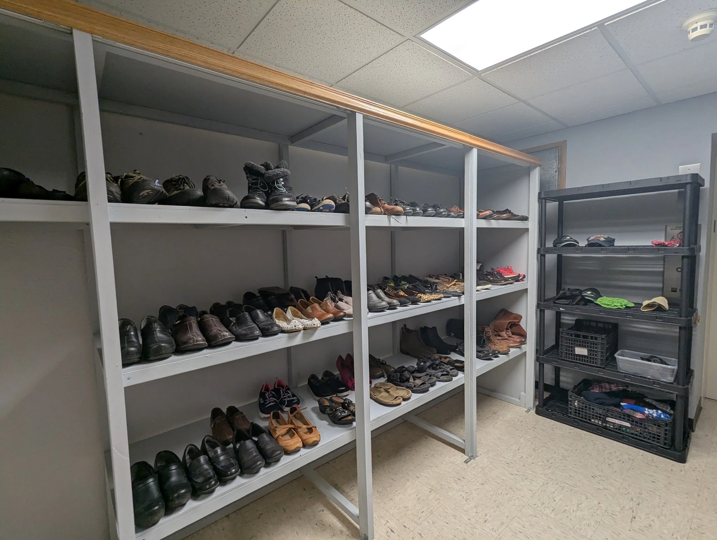 A room with metal shelves filled with various shoes, mainly black, brown, and tan, arranged on four levels. Some shelves are empty, and there is a black plastic crate with additional shoes on the right side of the image. The ceiling has white tiles, and the floor is covered with yellowish vinyl.