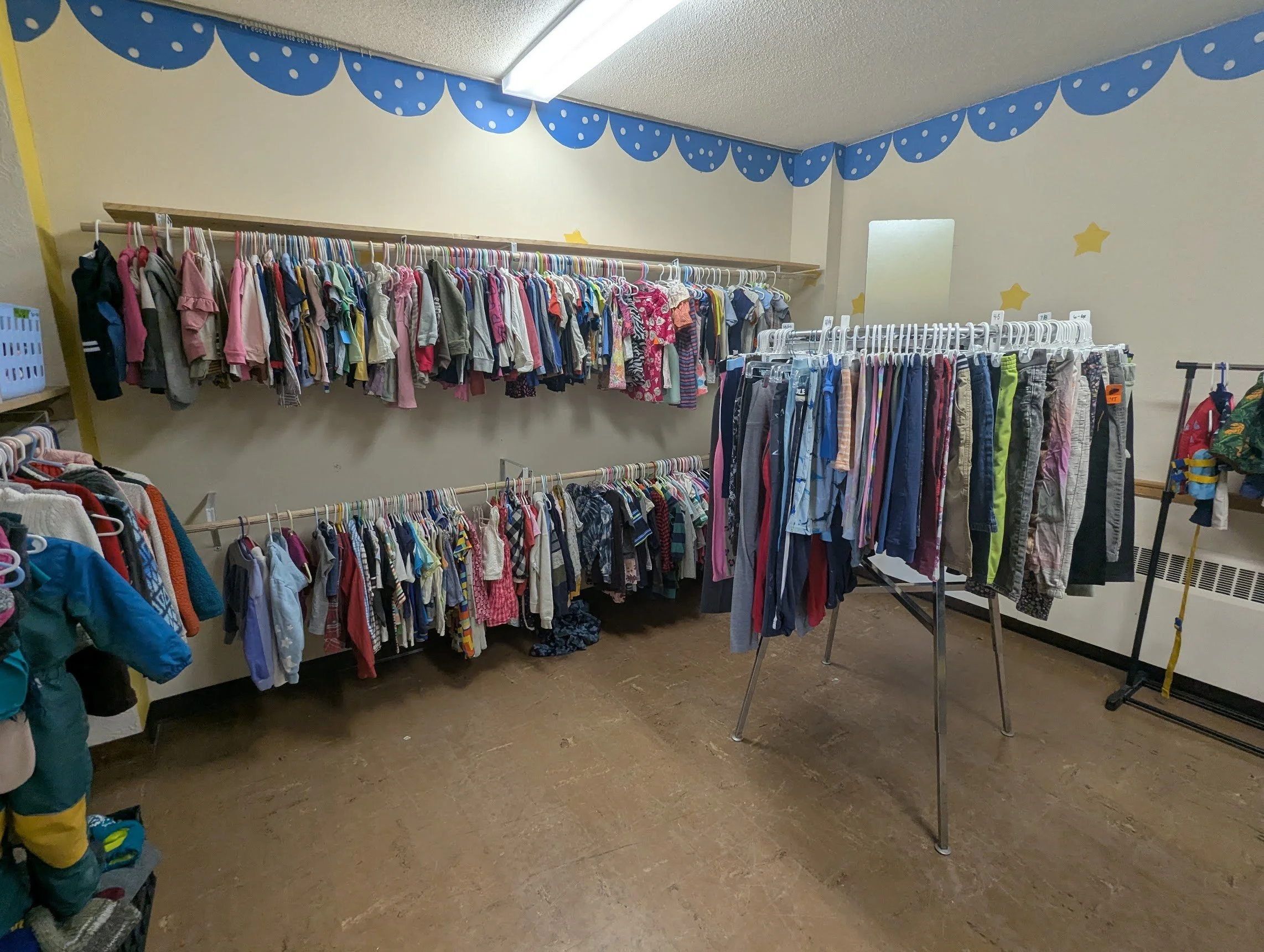 A room with walls decorated with blue scalloped border with white dots and yellow stars. Clothing items such as children’s shirts, pants, and jackets are hanging on white racks and hooks.